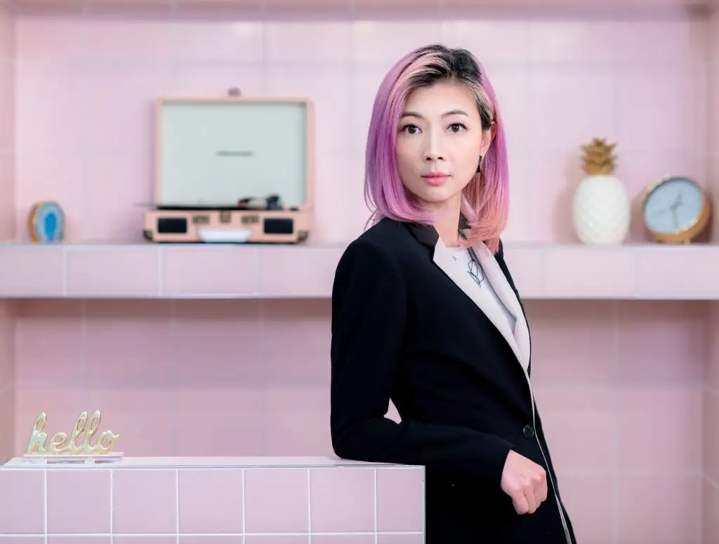 Interior Design - A woman with pink hair wearing a black suit, standing beside a pink tiled wall with decorative items. Dexign Matter Studio