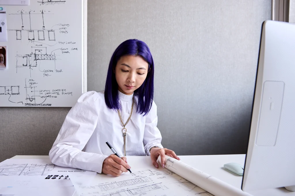 Interior Design - Interior designer with purple hair reviewing architectural plans at a desk with a computer and sketches. Dexign Matter Studio