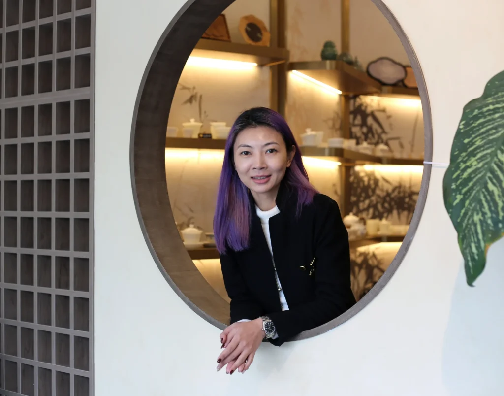 Interior Design - Portrait of Zoe-Lee with purple hair in a black blazer, leaning through a circular doorway in a modern interior with warm backlit shelves and minimalist decor. Dexign Matter Studio