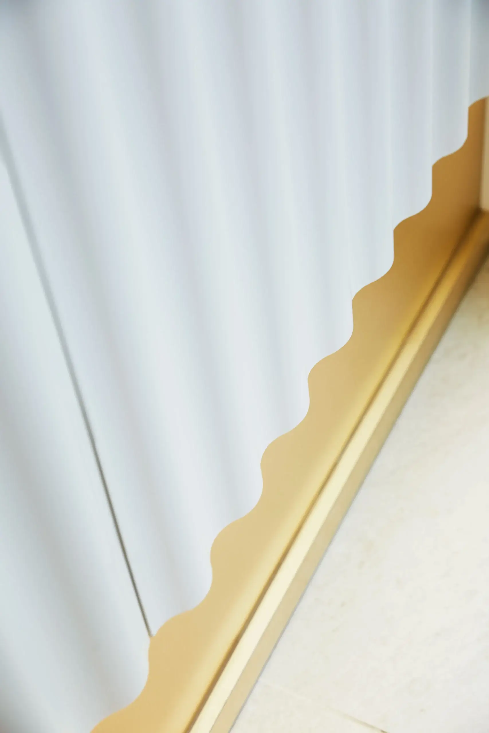 Close-up of a white curtain with a scalloped beige border along a wooden or tiled floor edge, creating a wavy pattern.
