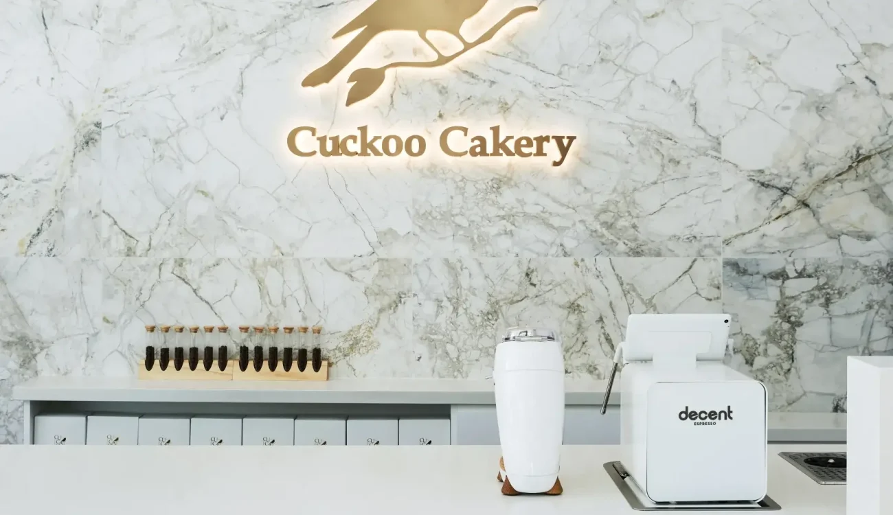 A marble-tiled wall backdrop with a glowing sign of a bird and the words 'Cuckoo Cakery' above a modern white counter. On the counter, a white coffee grinder and a small white blender are visible, with a row of small drawers in the background.