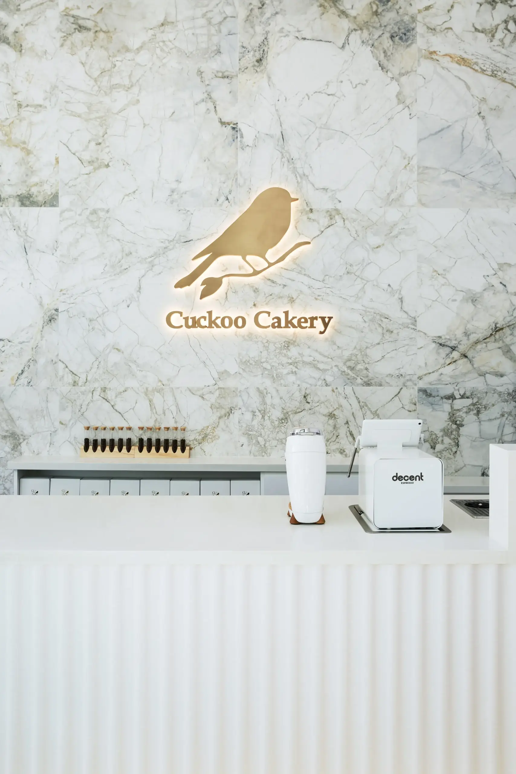 A marble-tiled wall backdrop with a glowing sign of a bird and the words 'Cuckoo Cakery' above a modern white counter. On the counter, a white coffee grinder and a small white blender are visible, with a row of small drawers in the background.
