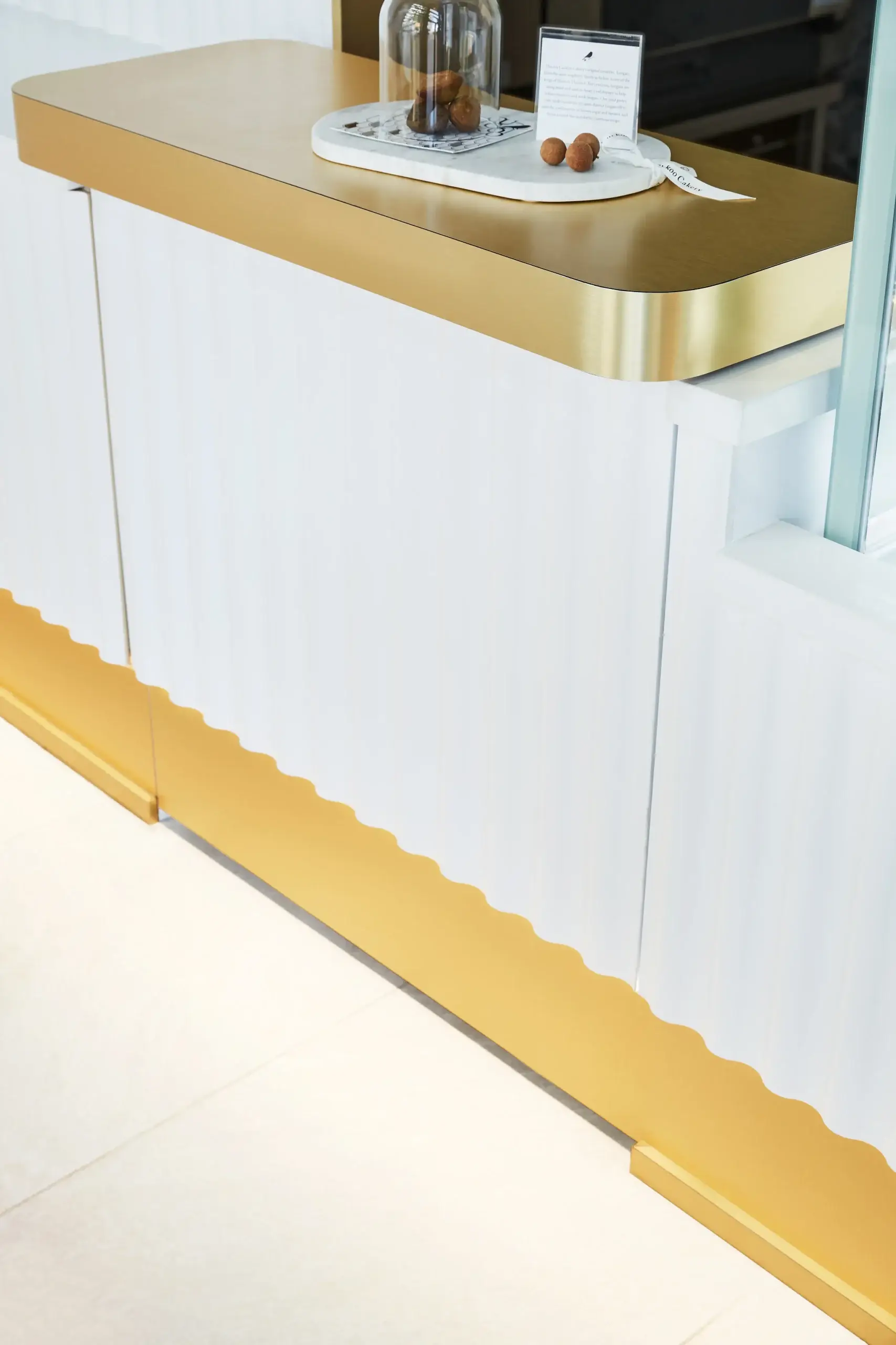 Stylish white reception desk with a wide gold countertop edge and a wavy gold trim along the bottom. On top sits a glass cloche and small display items, creating a luxurious showroom vibe.
