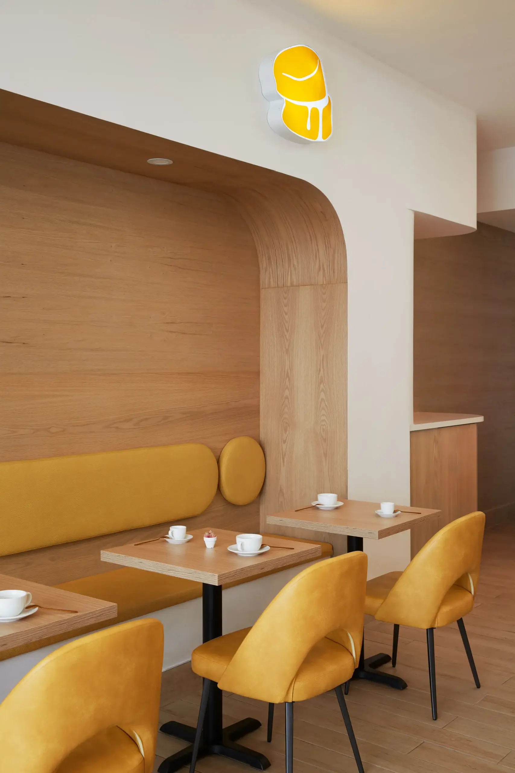 Interior of a modern cafe featuring mustard yellow chairs and a long cushioned bench along a wooden curved wall, small square tables set with cups and saucers, and a bright yellow logo on the white wall above.