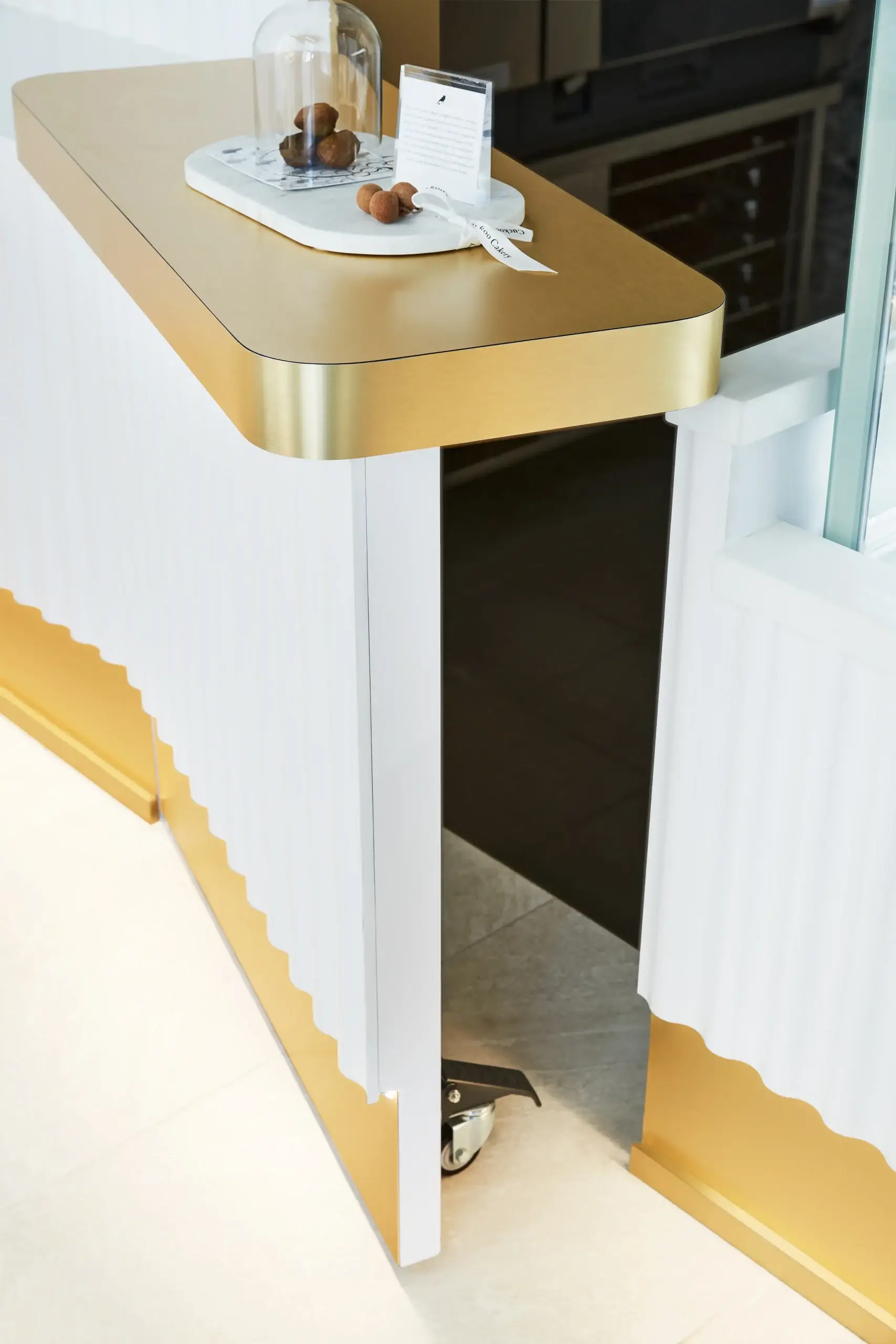 A stylish reception counter with a gold top and base trim. White paneling with scalloped gold accents sits near a glass window, and a small display with brown pastries is on the counter.