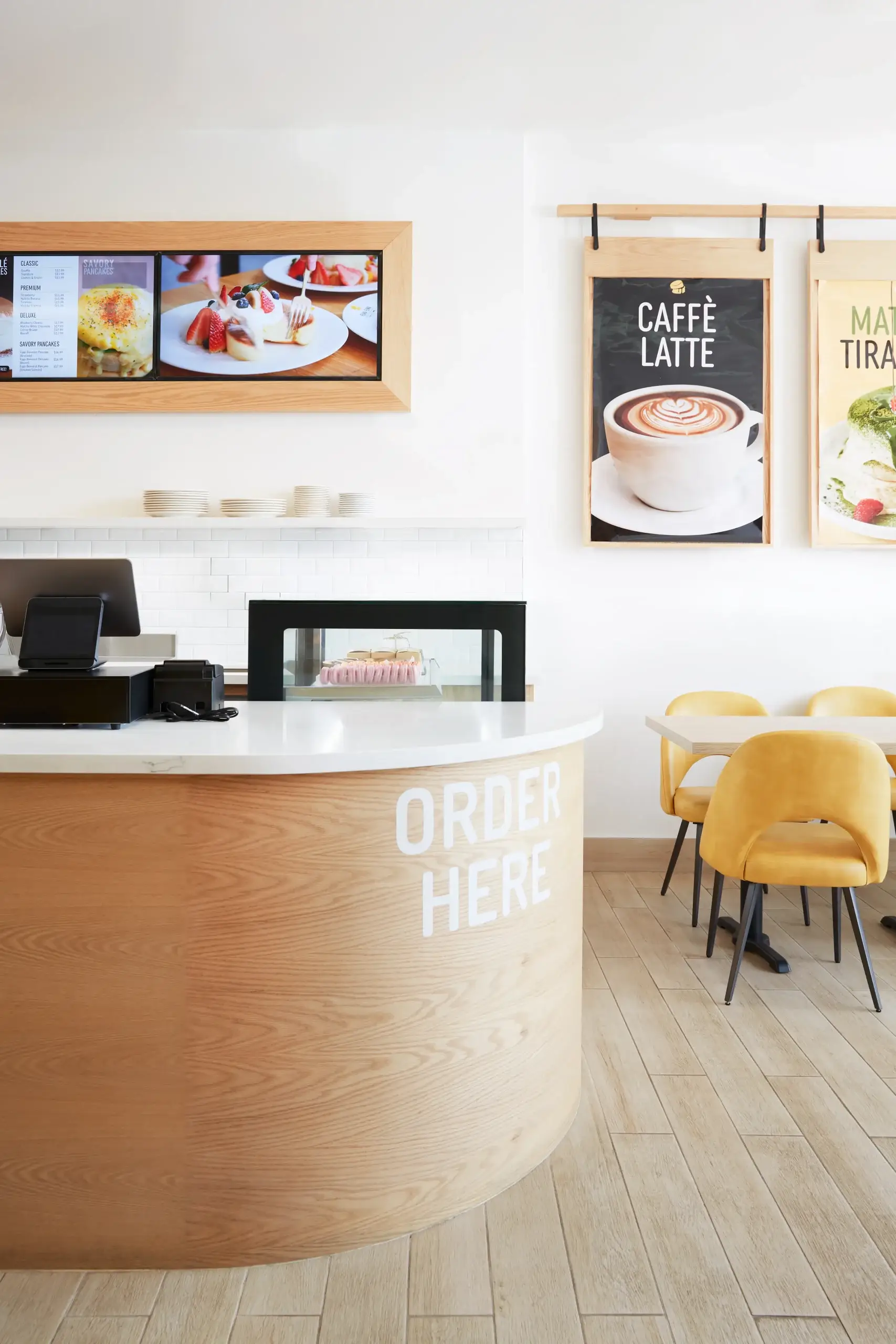 A modern, airy cafe interior featuring a curved wooden counter with the words ORDER HERE painted on it, a point-of-sale area, light wooden floors, and yellow chairs around light tables. Behind the counter are menu boards with images of desserts on the wall.