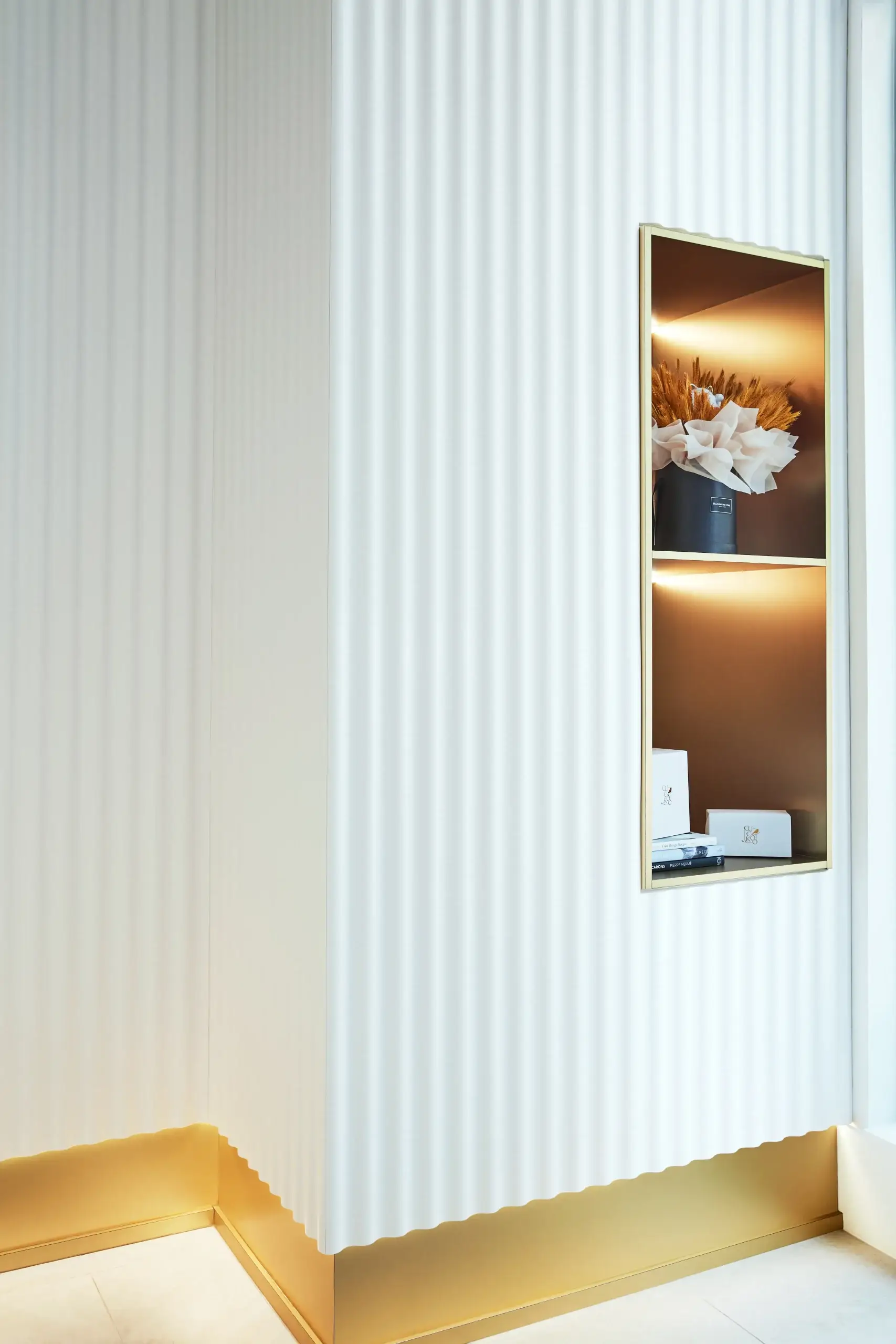 A tall white wall with vertical ribbed texture, featuring a slim rectangular framed display showing decorative items and warm lighting, set near a golden baseboard and light floor.