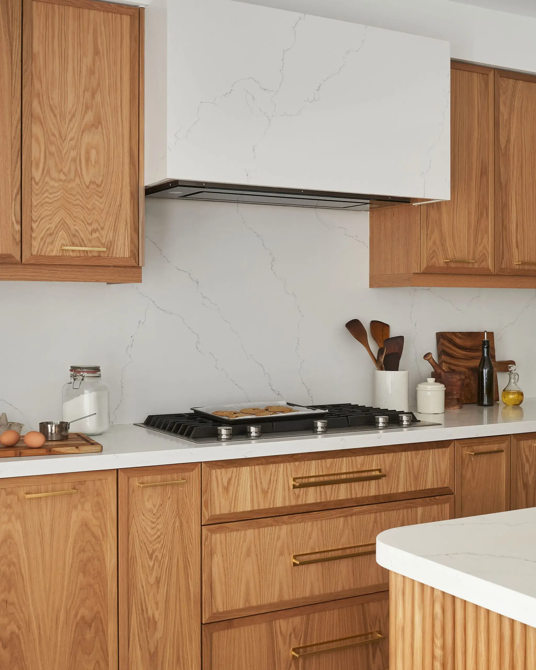 A modern kitchen featuring wooden cabinets with gold handles, a marble-veined white backsplash, and a gas stove built into a white countertop. Cooking utensils and bottles are arranged on the counter.