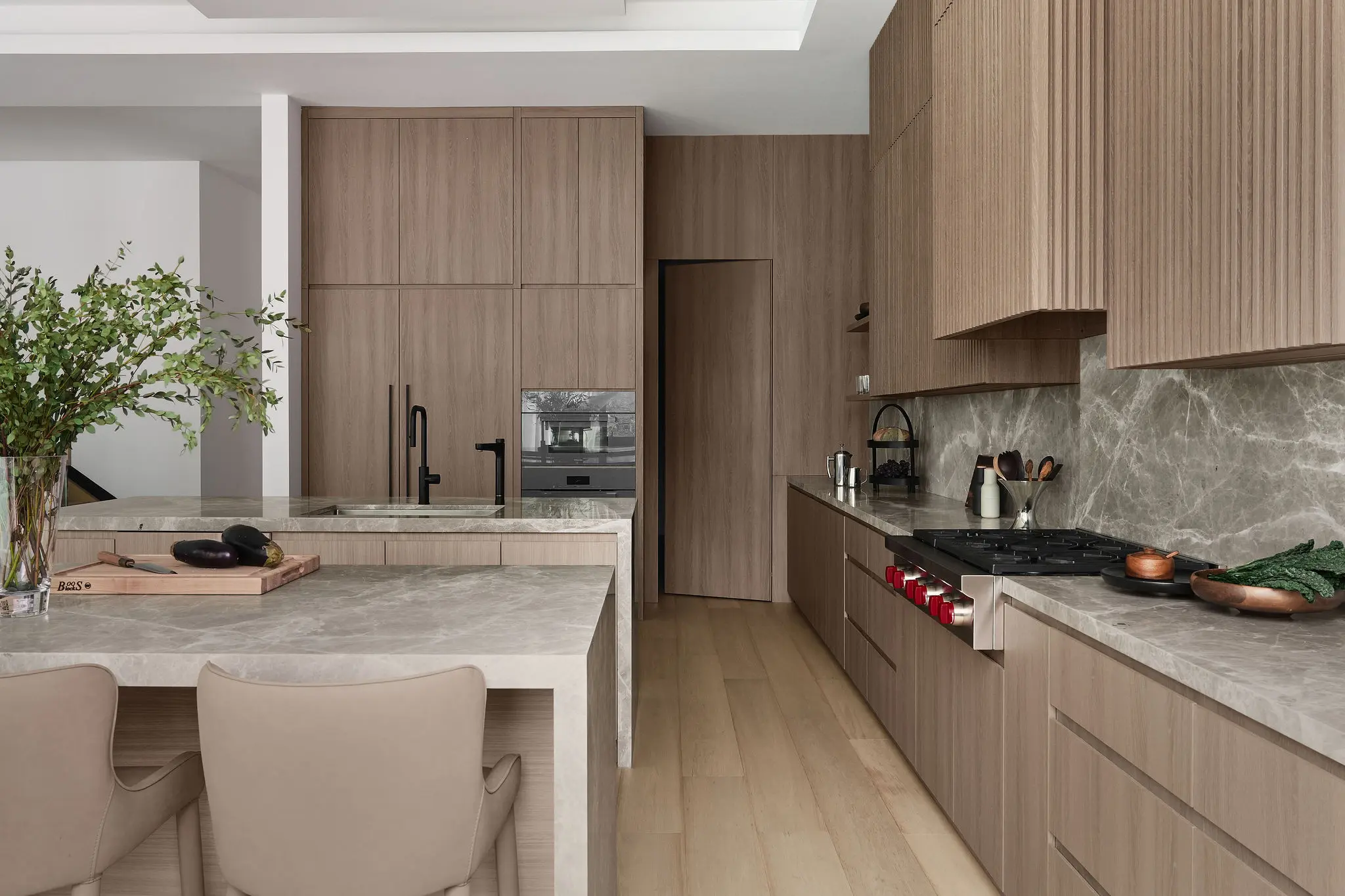 Warm-toned custom cabinetry and marble countertops form a streamlined kitchen, with a large leafy plant adding softness in a biophilic luxury interior design.