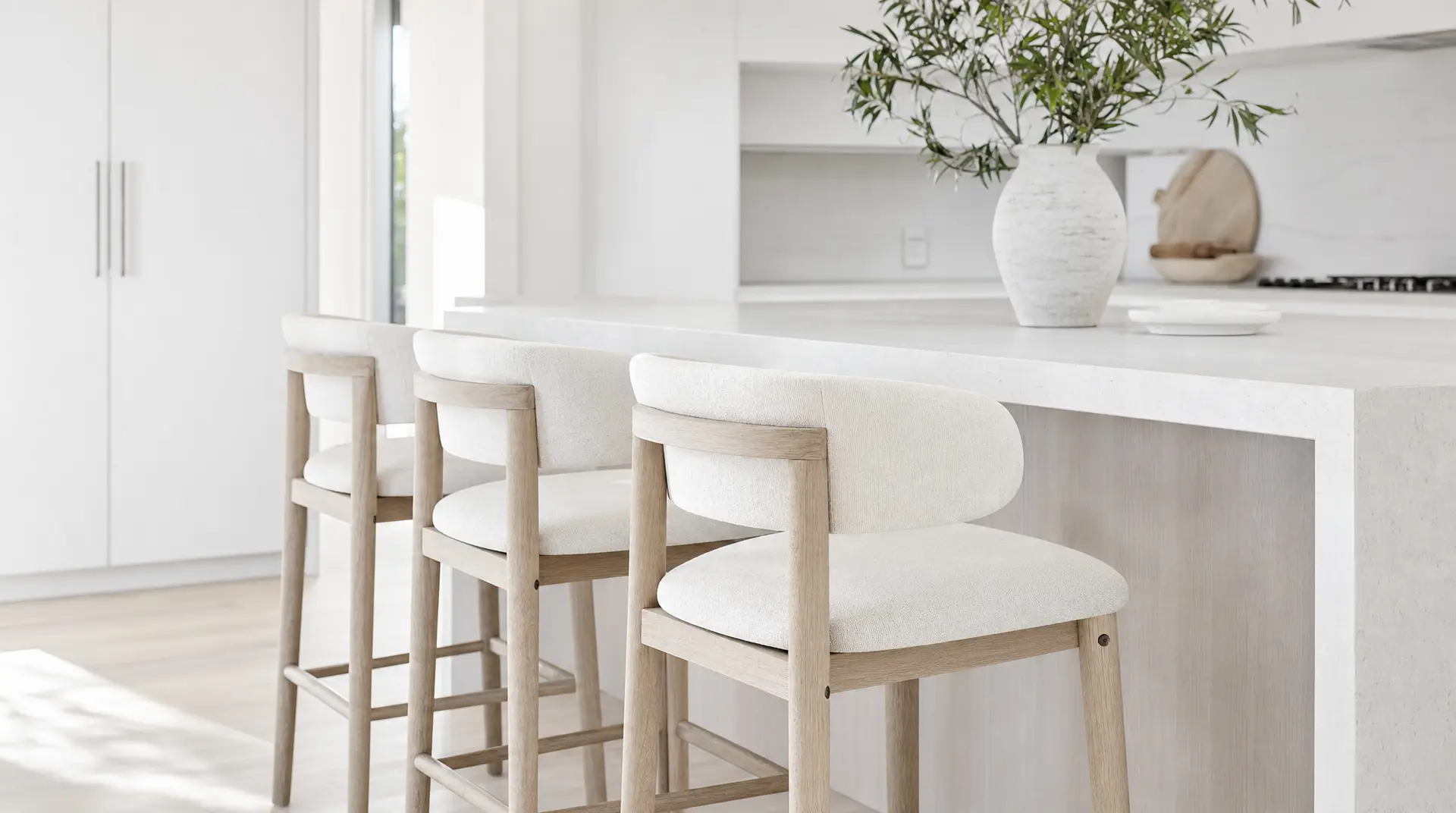 White dining counter with pale wood stools, leafy greenery in a ceramic vase, and bright daylight supporting maximizing natural residential light.