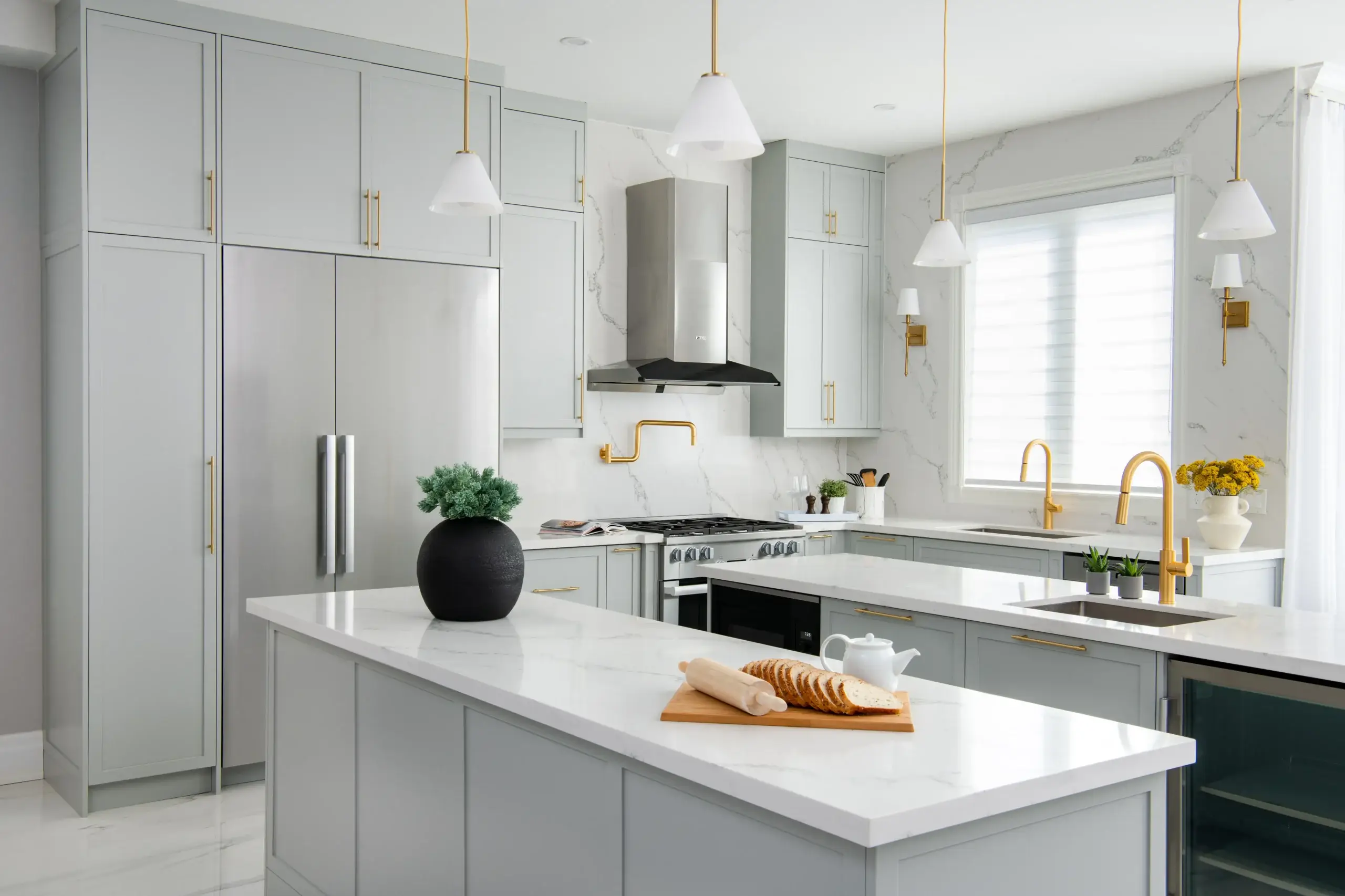 Light gray kitchen cabinets and a white marble island. Gold hardware and fixtures, including faucet and pendant lights, adorn the space. Stainless steel fridge and stove sit against marble-topped counters, with a loaf of bread on a cutting board and a black plant pot on the island.