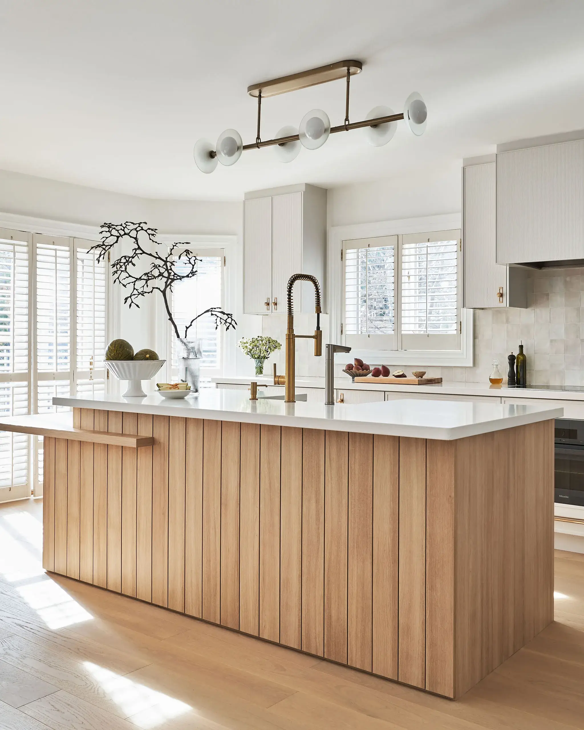 Bright kitchen with a large wooden paneled island, white countertop, brass faucet, and white cabinetry. Sunlight streams through shutters, modern chandelier above, and decorative plants and fruit on the counter.