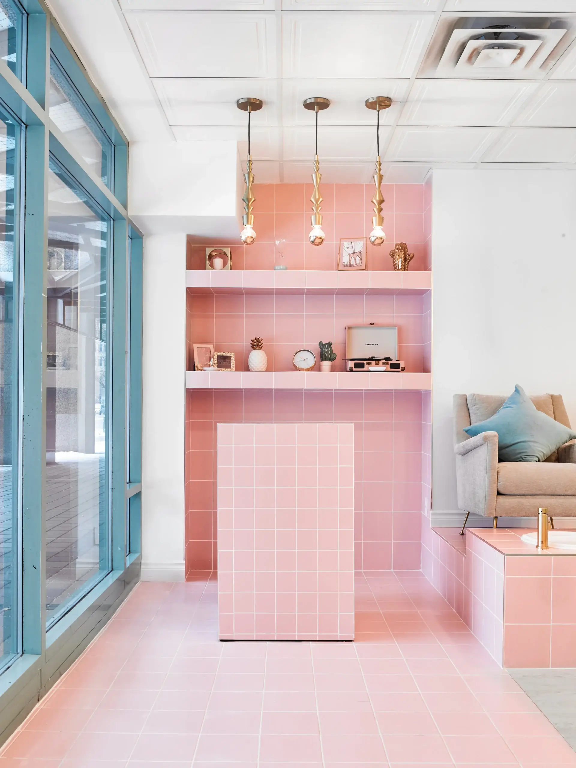 A bright interior features pink tiled walls and floor, a pink tiled island, and shelves with retro decor. A blue glass door wall runs along the left, while a beige chair with blue cushion sits to the right. Three hanging light fixtures with gold accents dangle from the white ceiling.