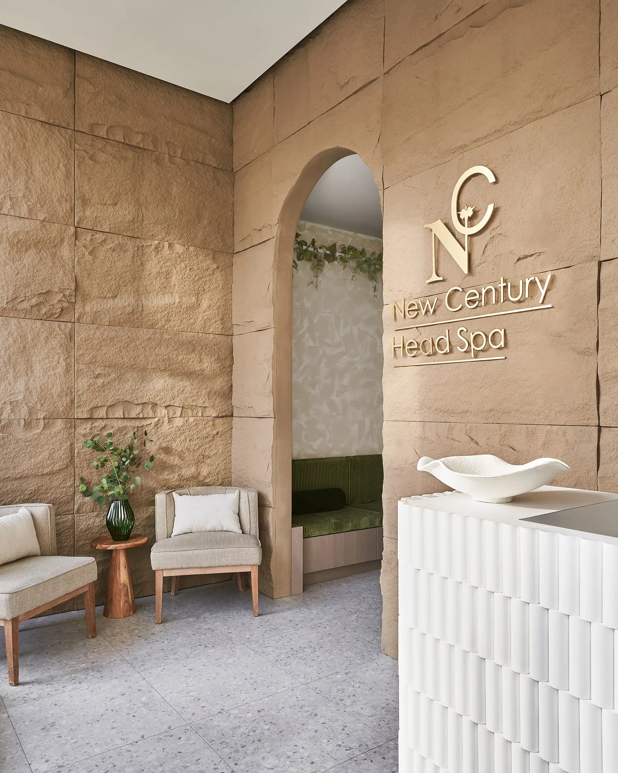 Spa reception area with beige stone walls, arched doorway, and a sign reading 'New Century Head Spa'. Comfortable seating with beige chairs, a small wooden side table with a plant, and a white textured reception counter in the foreground.