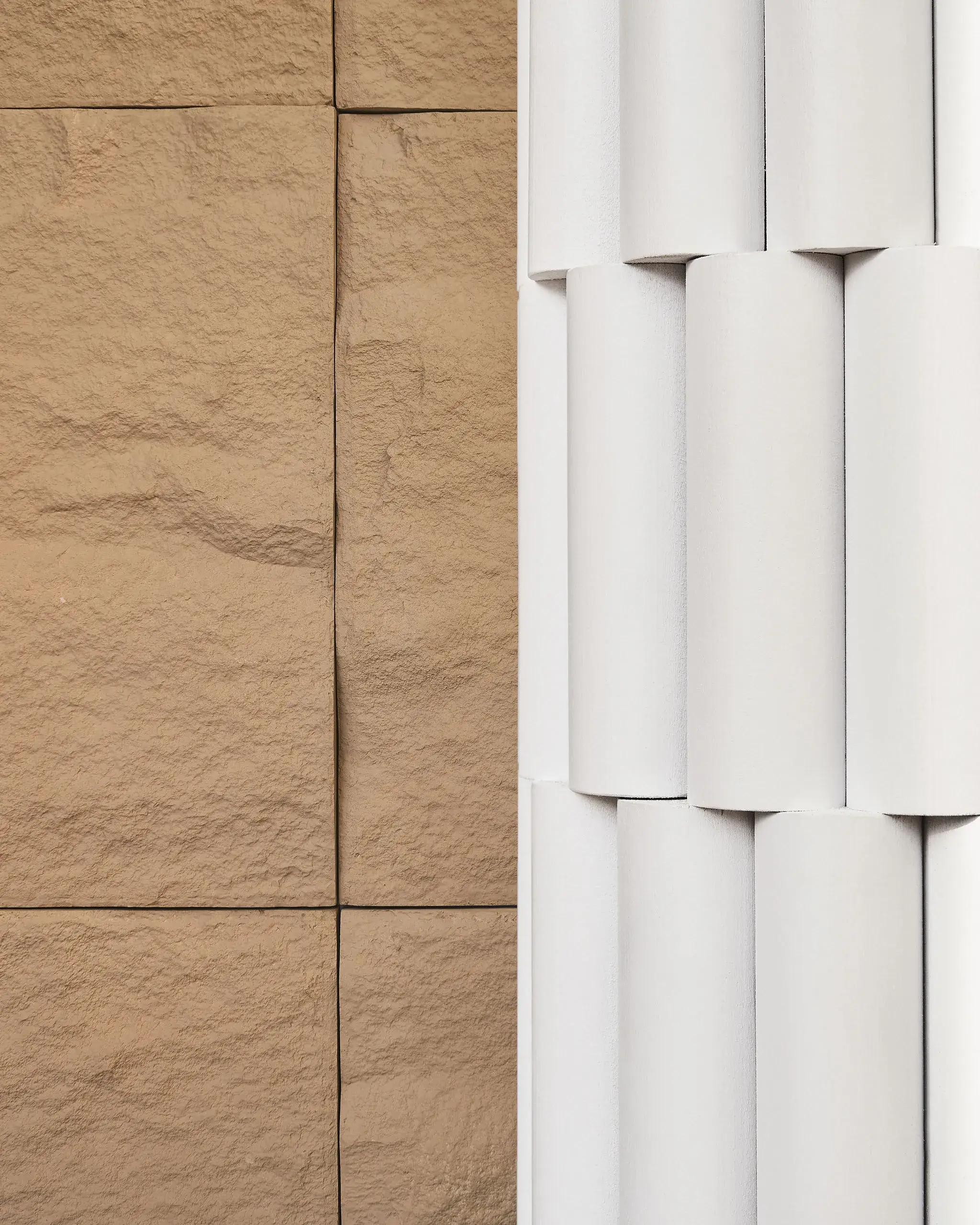 A close-up of two architectural textures: on the left, brown textured stone blocks forming a flat wall. On the right, white cylindrical columns arranged in a neat vertical stack.
