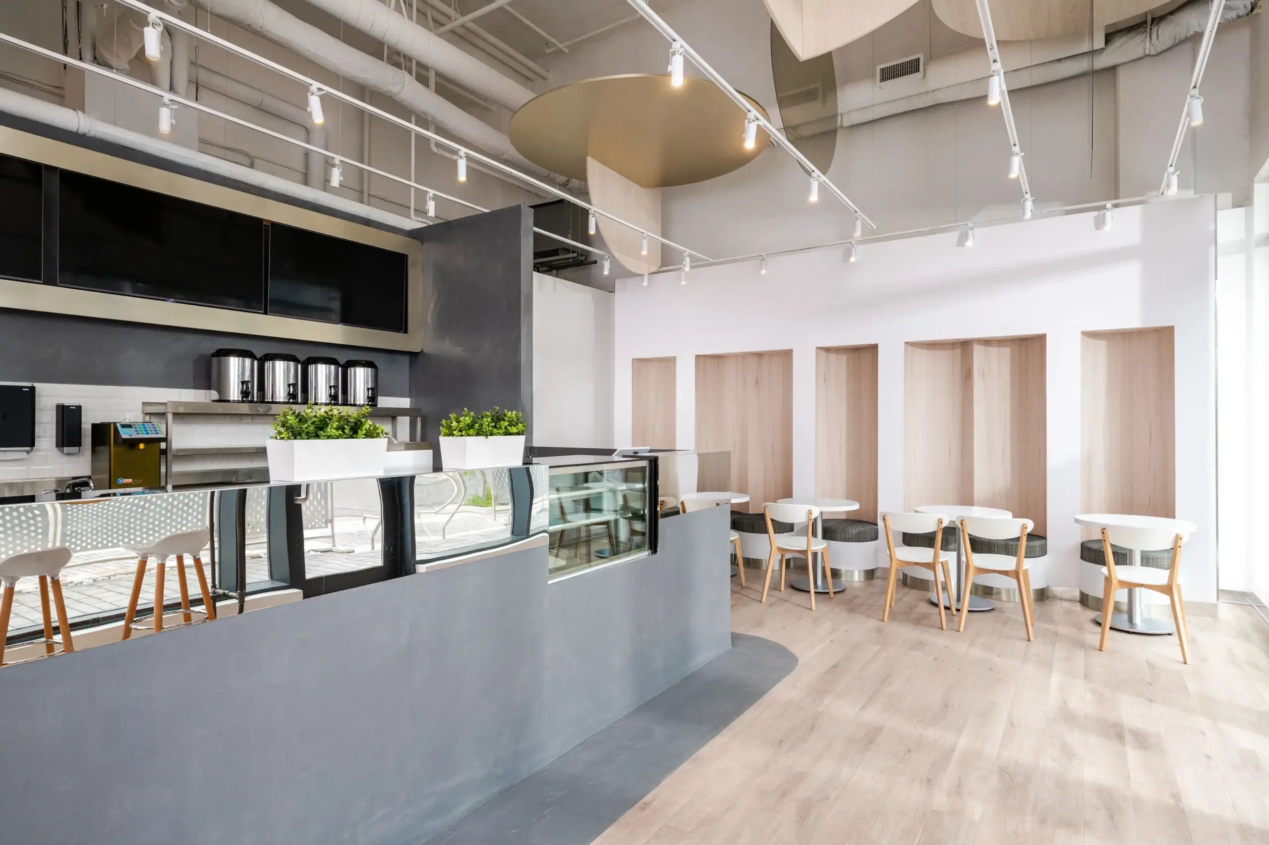 A sleek, contemporary cafe with a curved gray counter, glass display case, and bar-height seating. Light wood floors, white walls, and round white tables with wooden chairs line a wall with built-in wooden alcoves.