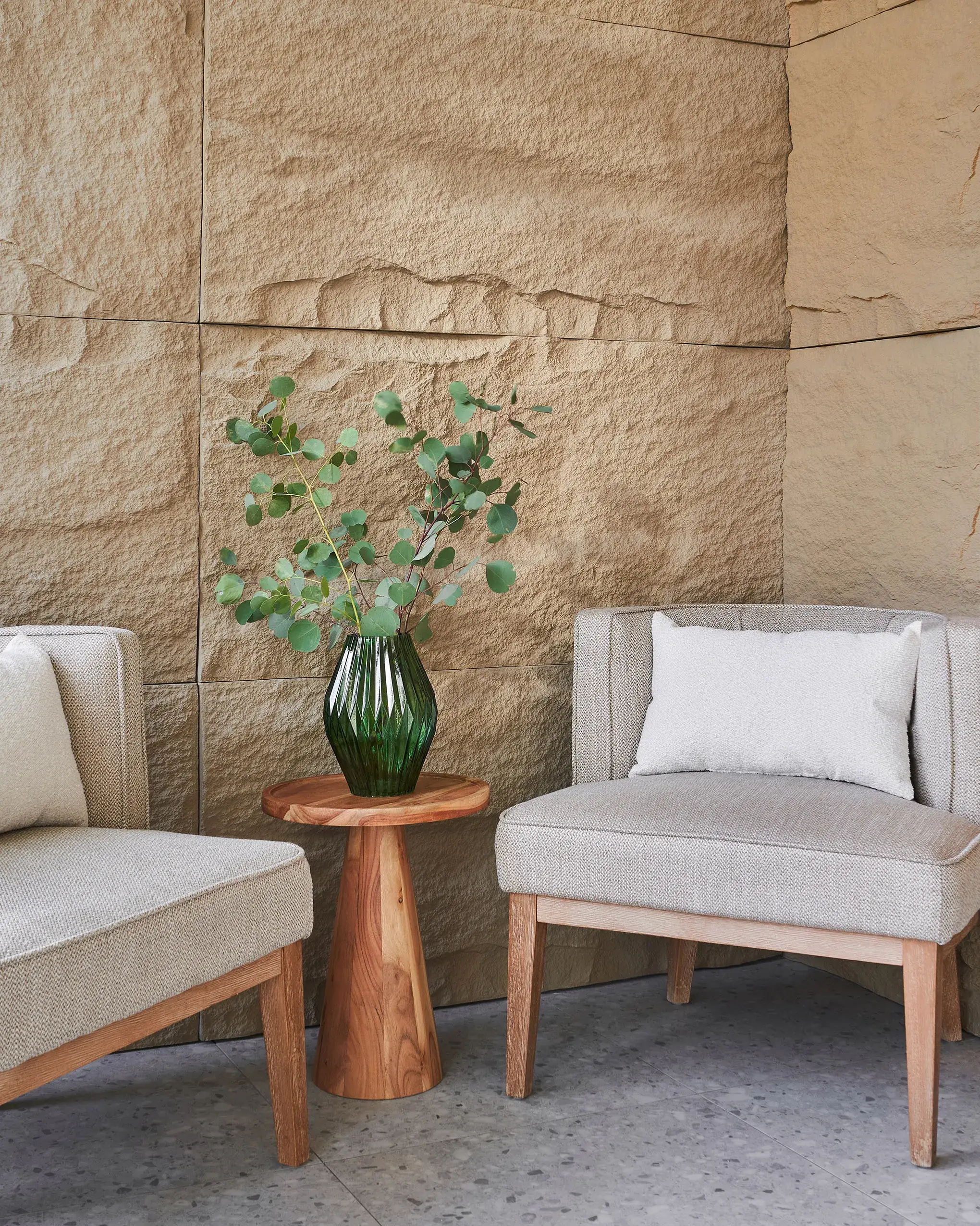 A small seating area against a textured beige stone wall, featuring two beige armchairs, a round wooden side table, and a dark green geometric vase with eucalyptus stems.