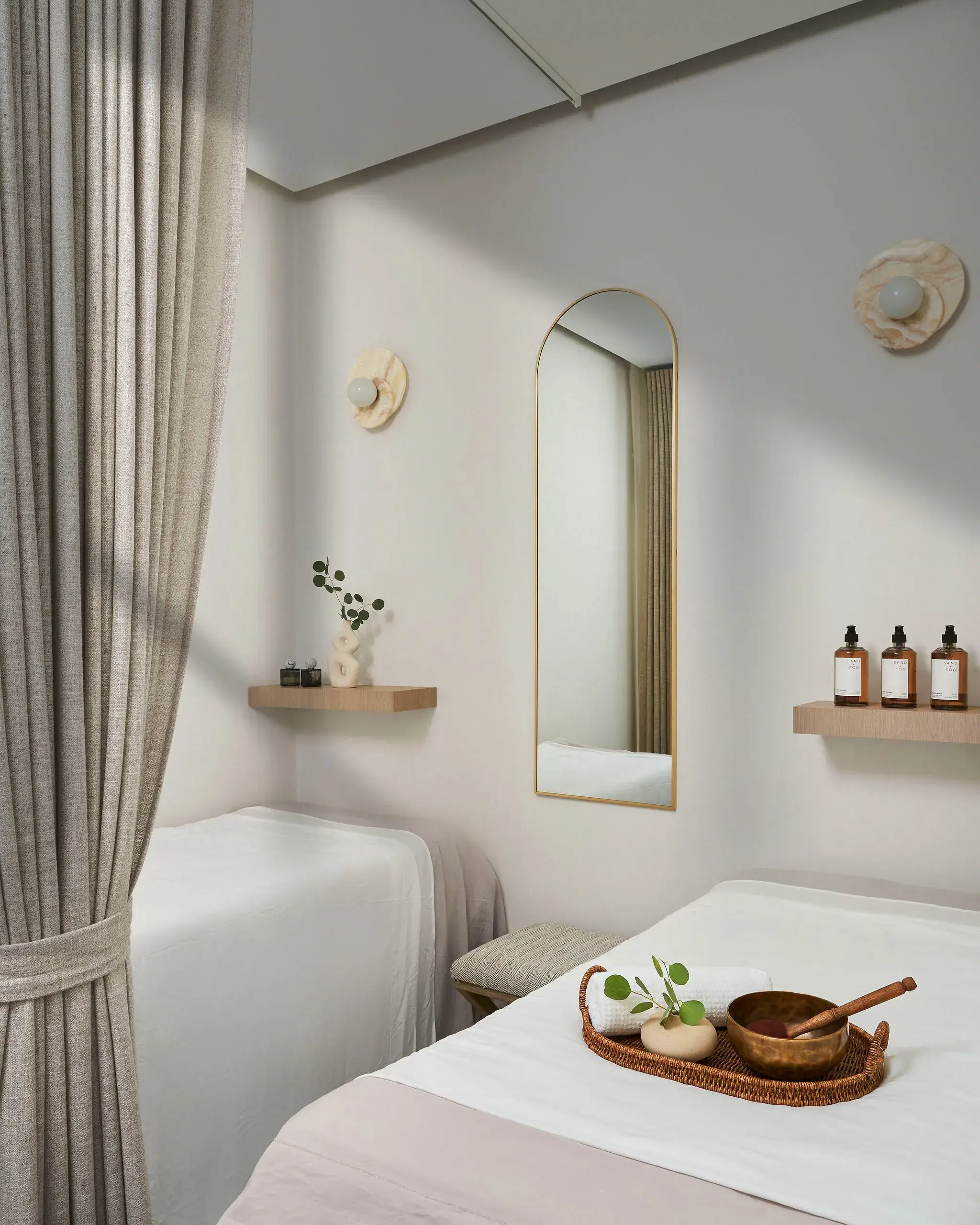 A serene spa room with two single beds, light beige curtains, and a tall arched mirror with a gold frame on a white wall. Small shelves hold bottles and decor, a woven tray with a wooden ladle and greenery rests on a bed, creating a calm, neutral color palette.