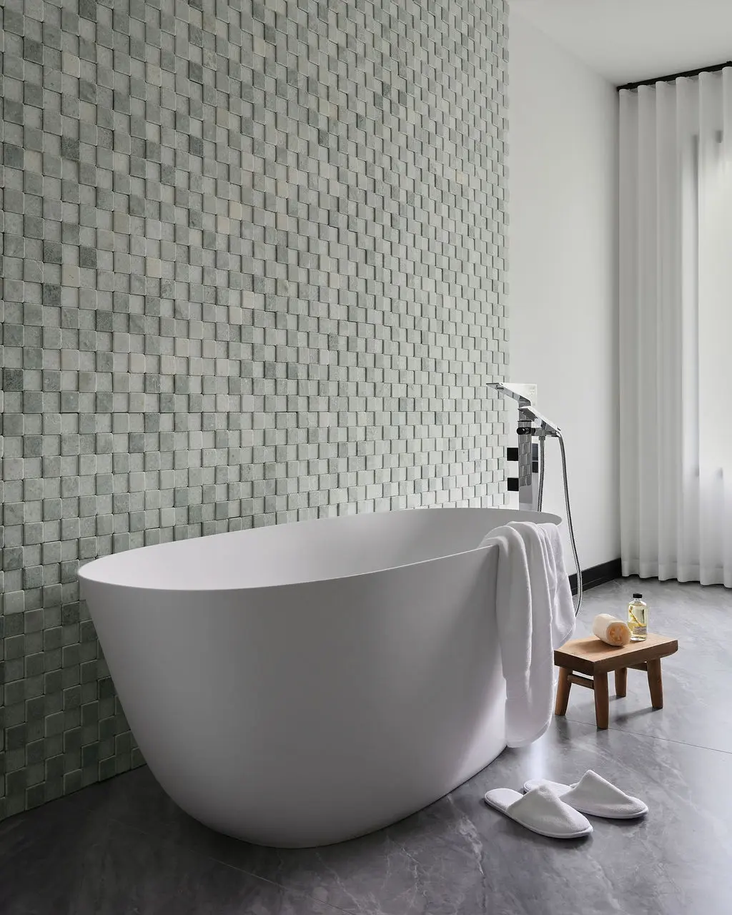 Interior Design - A freestanding white bathtub sits beside a textured gray tile wall in a minimalist bathroom with a shower, towel, and stool. Dexign Matter Studio