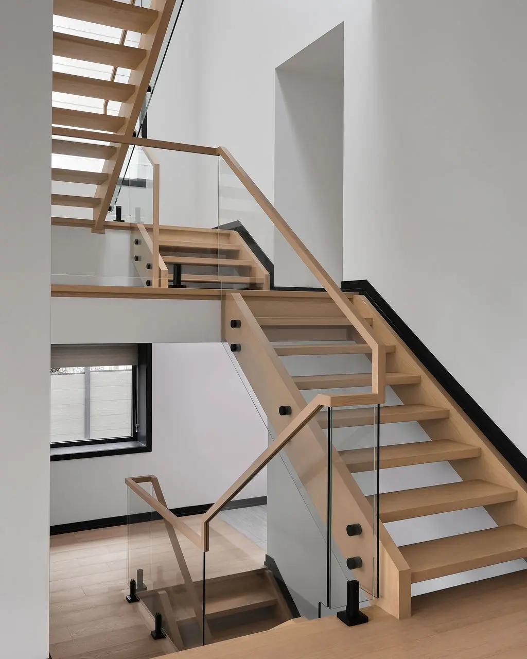 Interior Design - A modern staircase with light wood treads, glass railings, and black trim rises through a bright white interior. Dexign Matter Studio