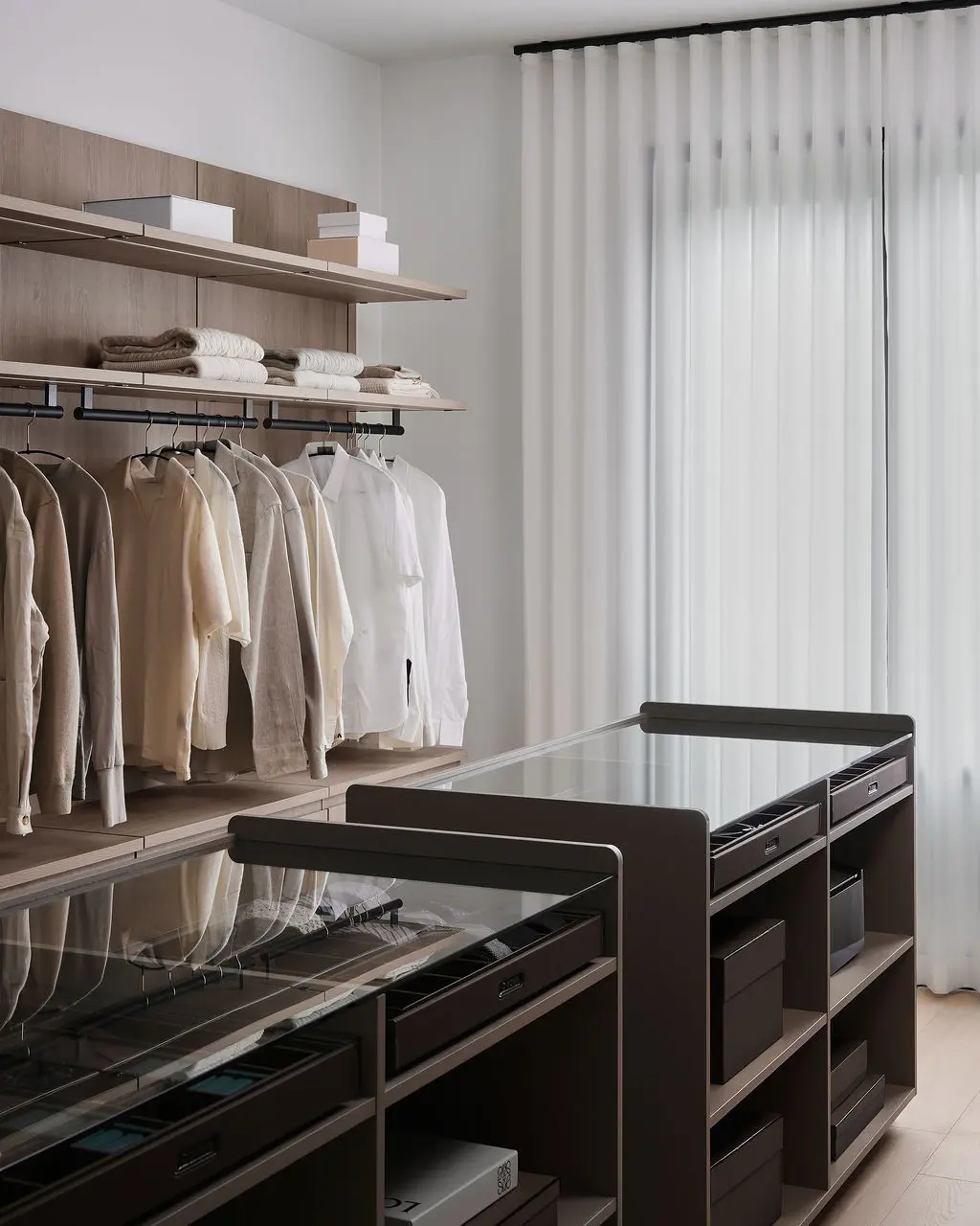Interior Design - A walk-in closet with wood shelving, hanging neutral clothing, folded linens, and dark glass-topped storage islands. Dexign Matter Studio