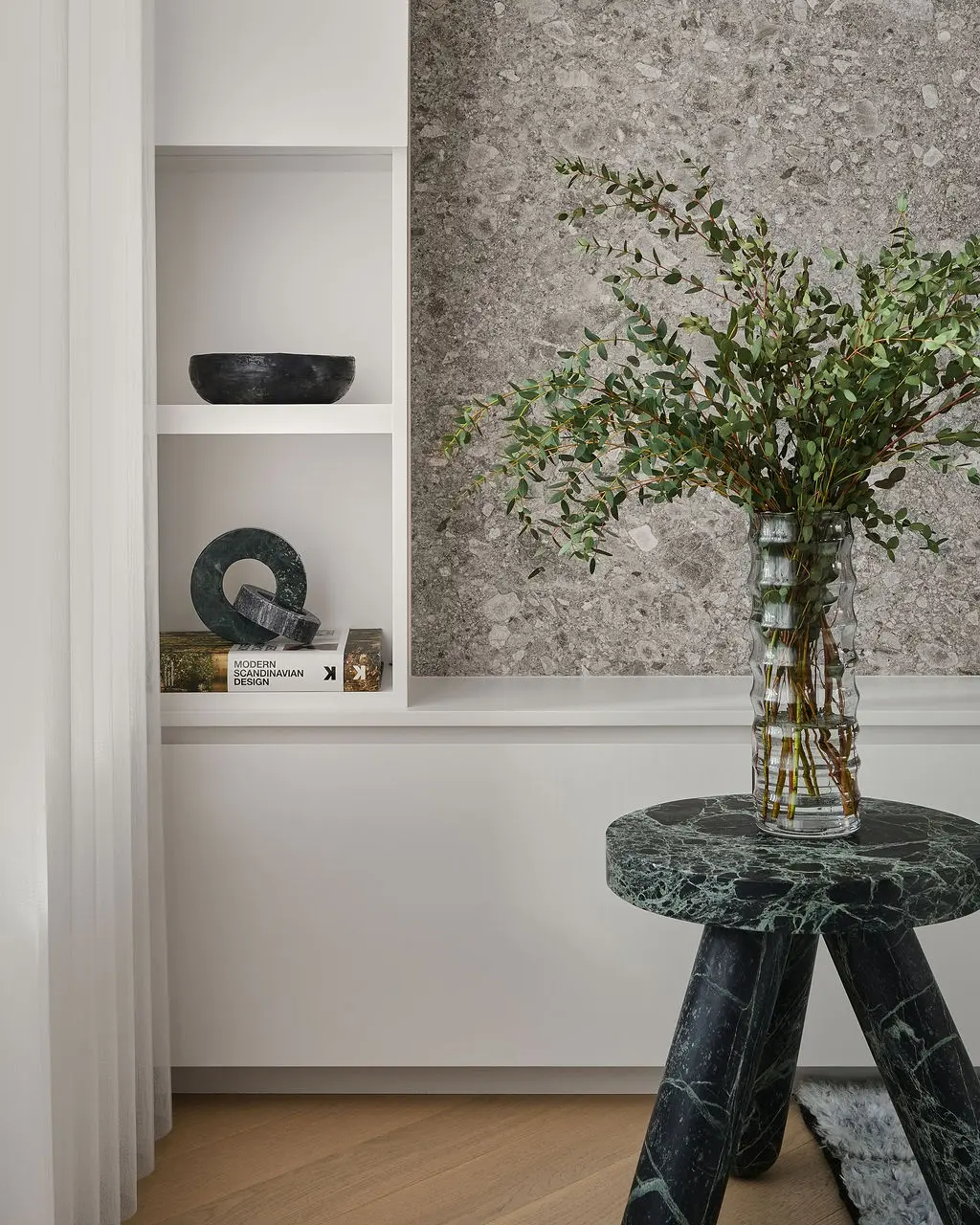 Interior Design - A glass vase of leafy branches sits on a green marble side table beside built-in shelves and sculptural decor. Dexign Matter Studio