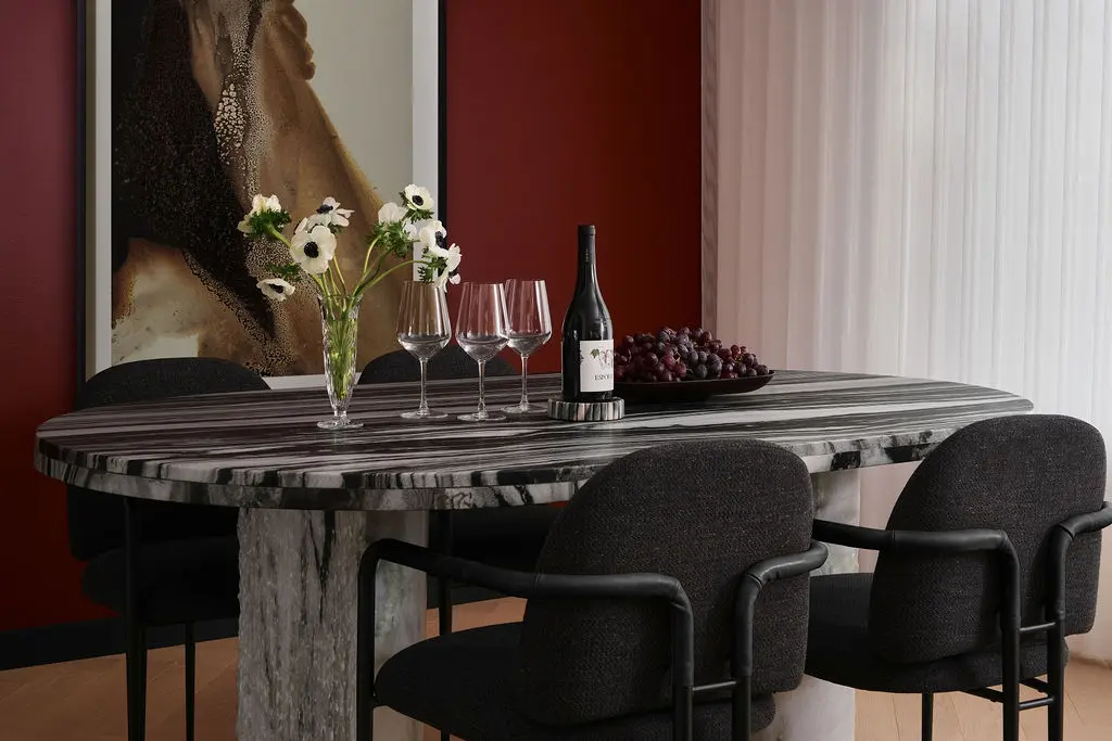 Interior Design - An oval marble dining table sits beside a red wall, with dark chairs, wine glasses, a bottle, white flowers, and grapes. Dexign Matter Studio