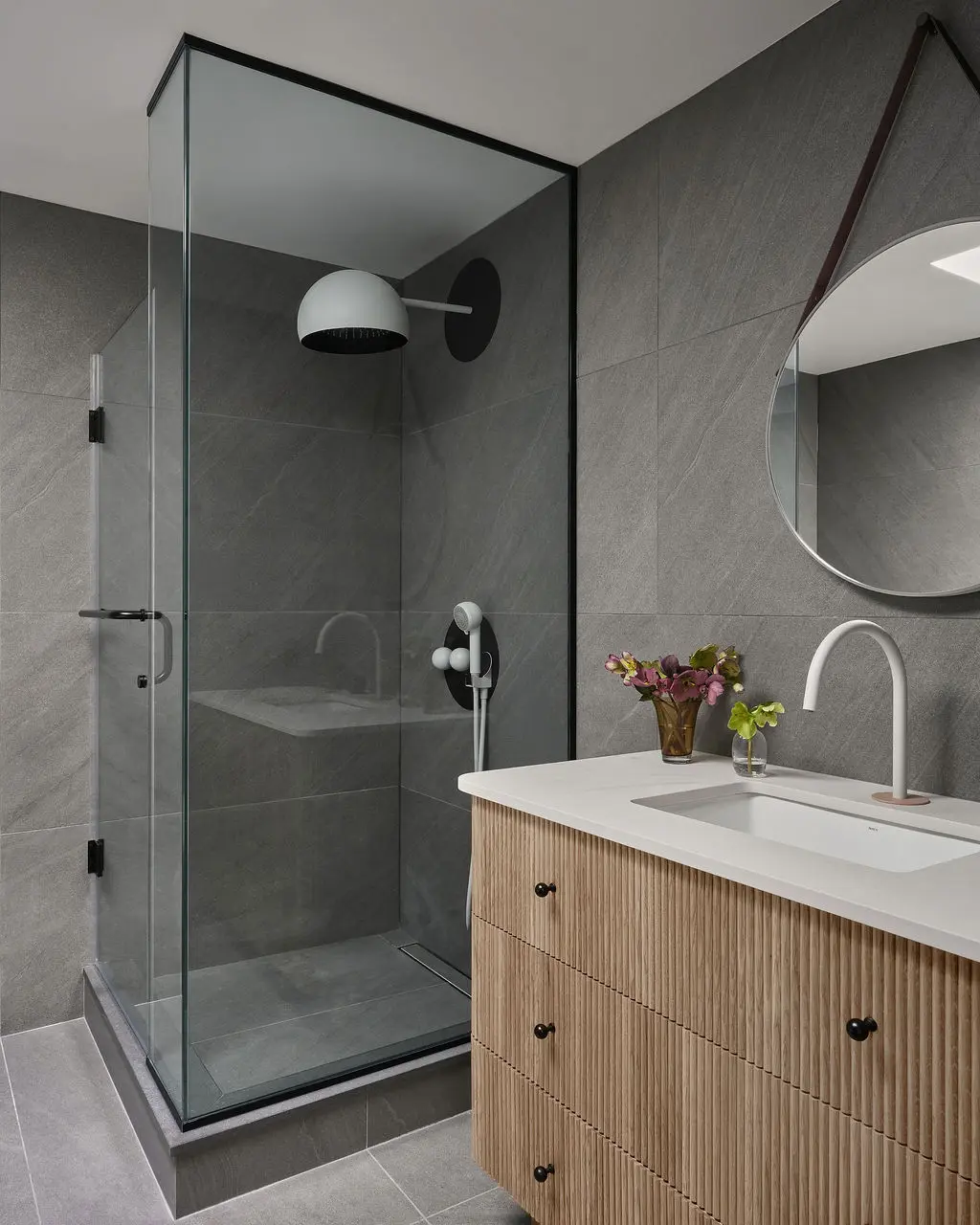 Interior Design - A gray tiled bathroom features a glass shower, round mirror, wood vanity, and white countertop sink. Dexign Matter Studio