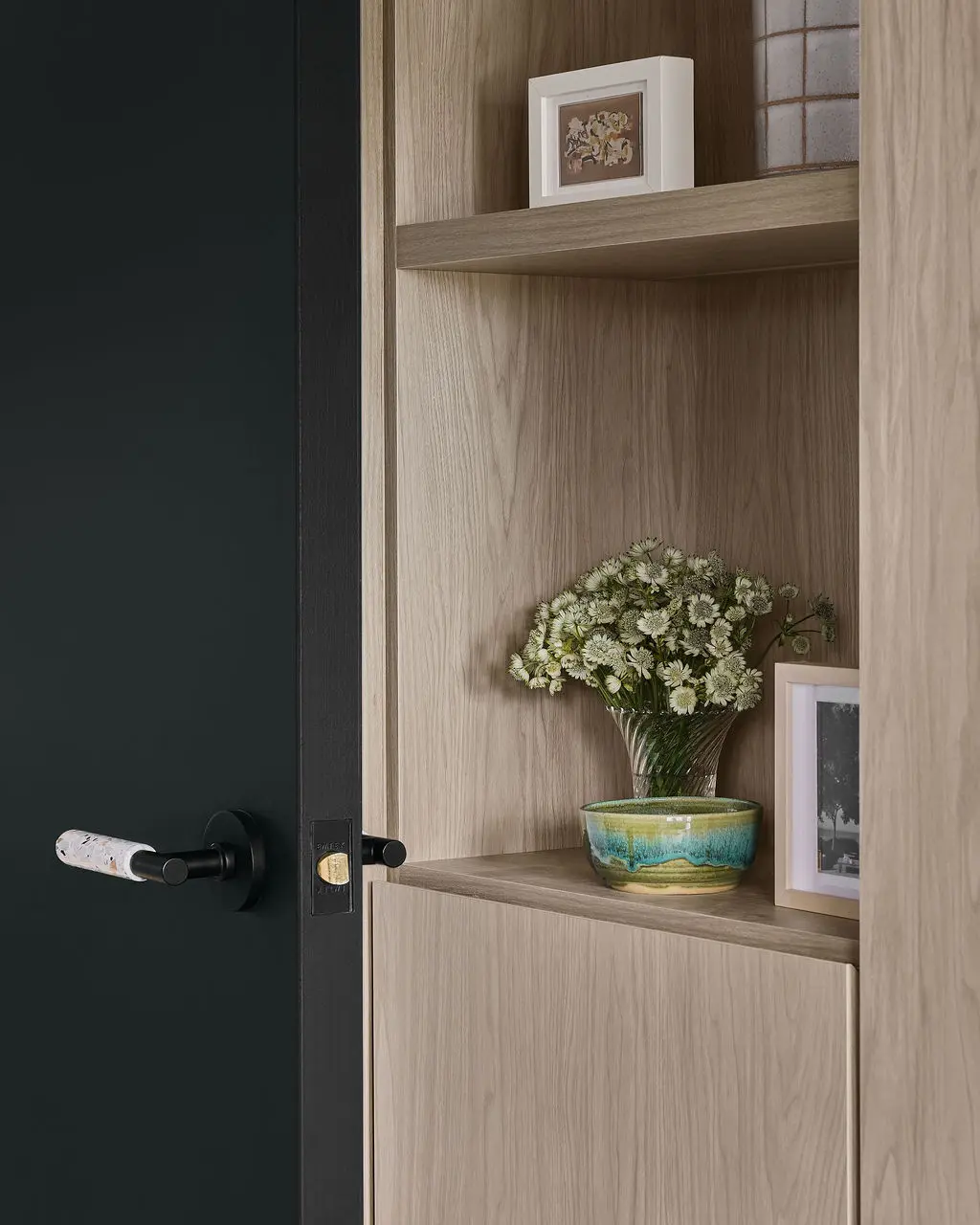 Interior Design - A dark green door opens beside a light wood shelf niche holding flowers, framed art, and a glazed ceramic bowl. Dexign Matter Studio