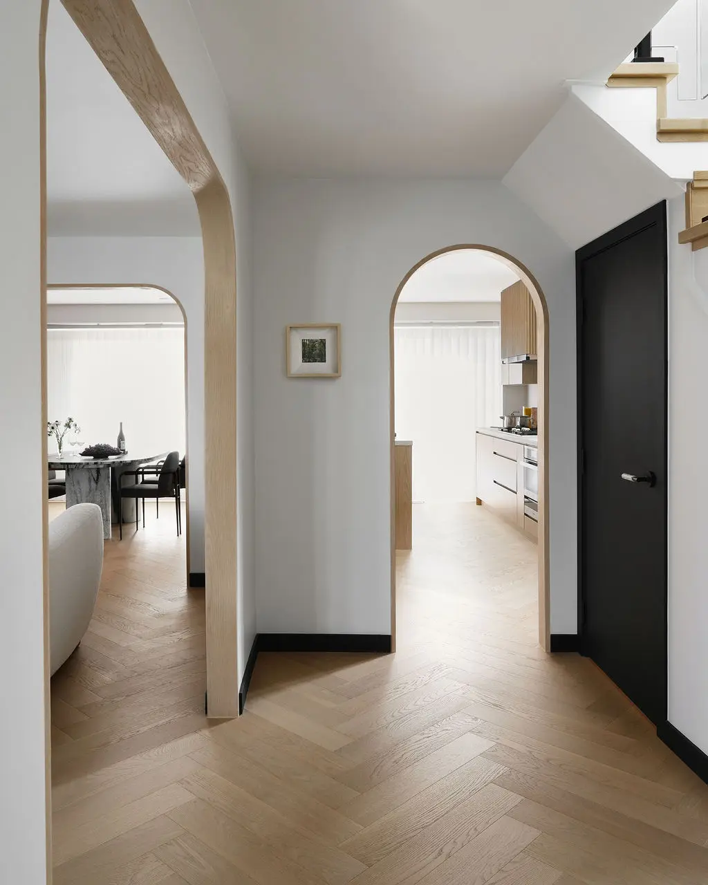 Interior Design - A light hallway with arched openings leads to a dining area and a bright kitchen, with herringbone wood flooring and a black door. Dexign Matter Studio