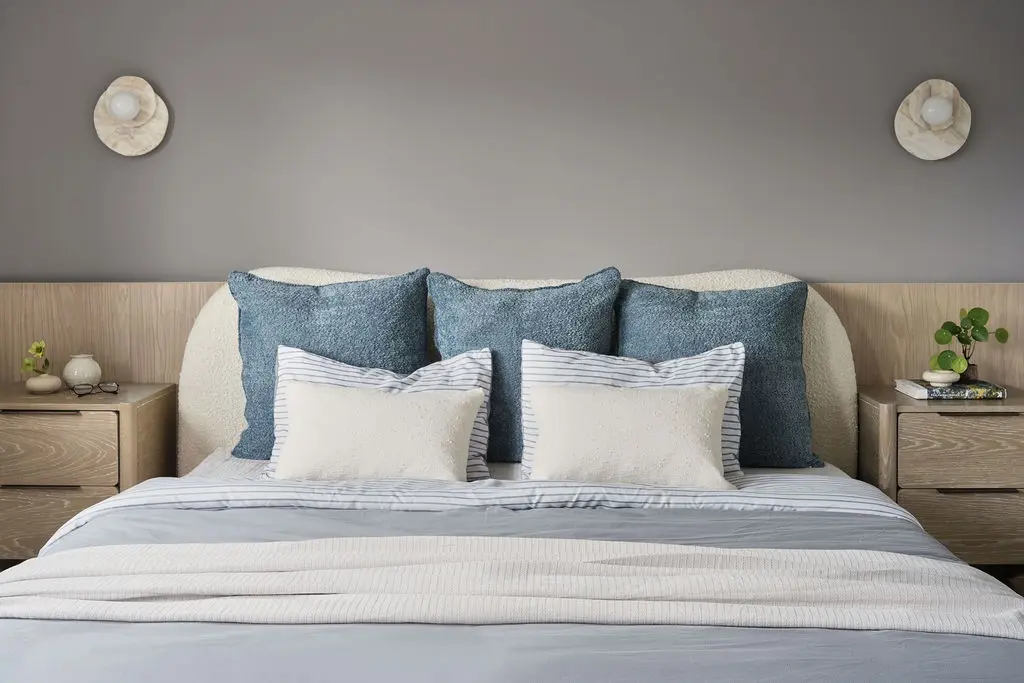 Interior Design - A neatly made bed with blue cushions and striped pillows sits against a gray wall with matching sconces and wood nightstands. Dexign Matter Studio