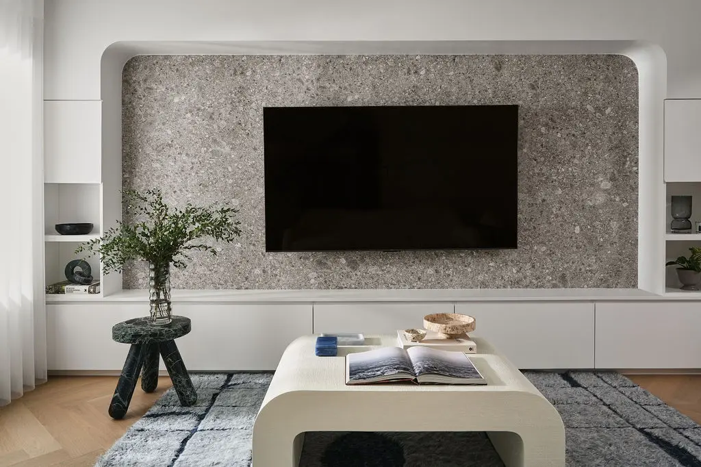 Interior Design - A modern living room features a stone-textured TV wall, white built-in cabinets, and a curved coffee table with an open book. Dexign Matter Studio