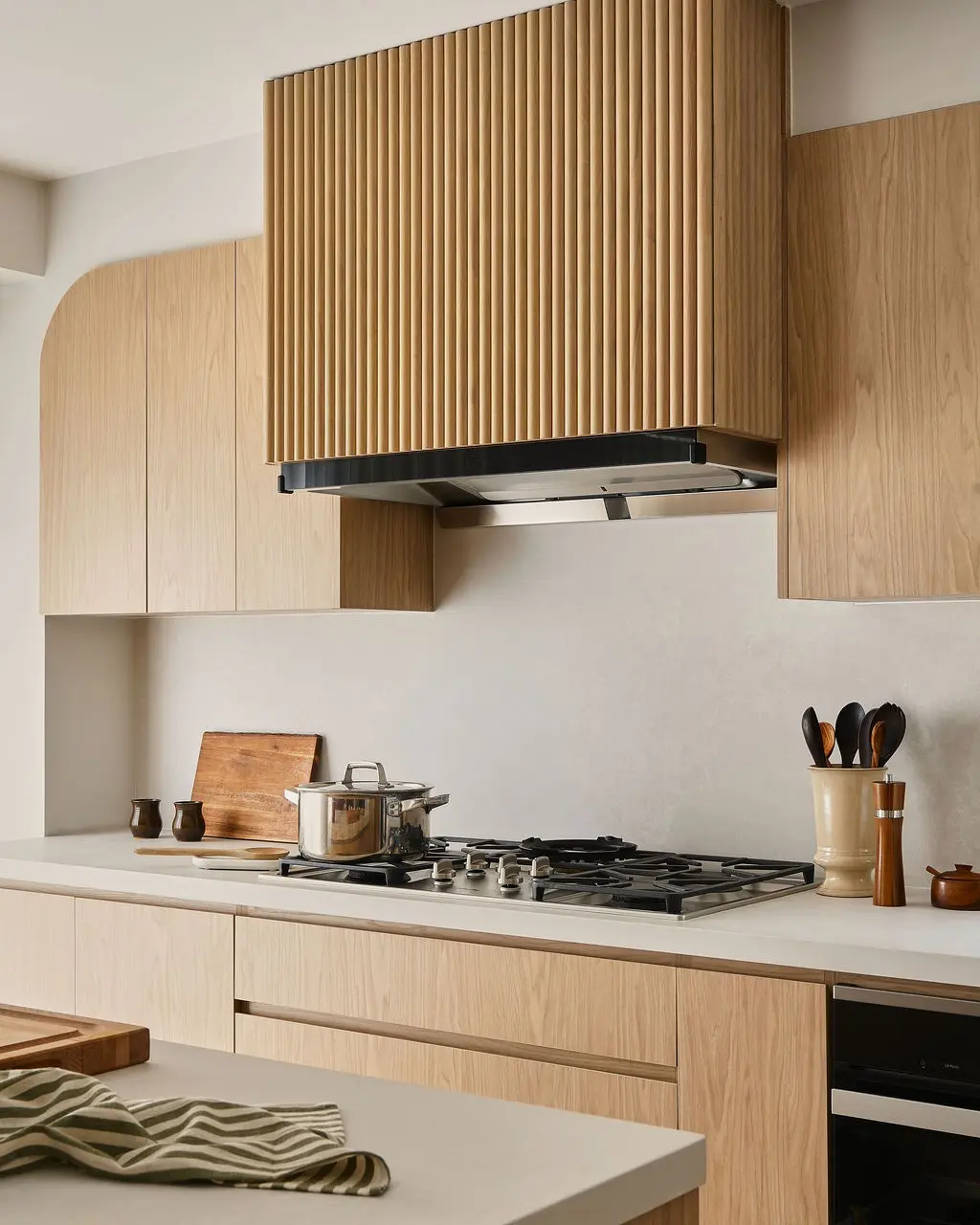 Interior Design - Light wood cabinetry and a ribbed range hood frame a minimalist kitchen with a gas cooktop, stainless pot, and pale counters. Dexign Matter Studio