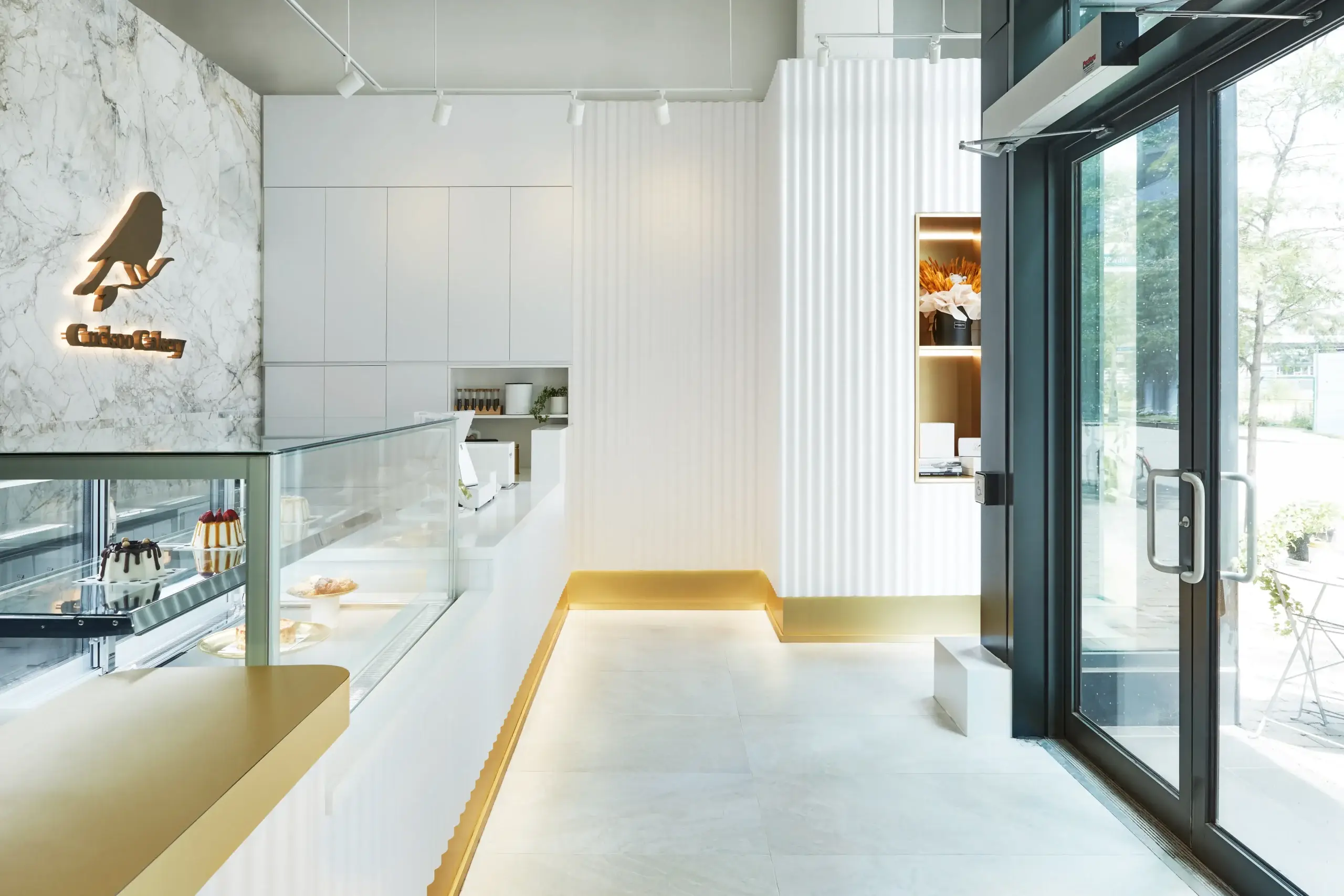 A sleek, bright bakery interior with white marble wall, glass display case showcasing cakes, and a gold-accented seating ledge. Large glass doors let in natural light from outside.