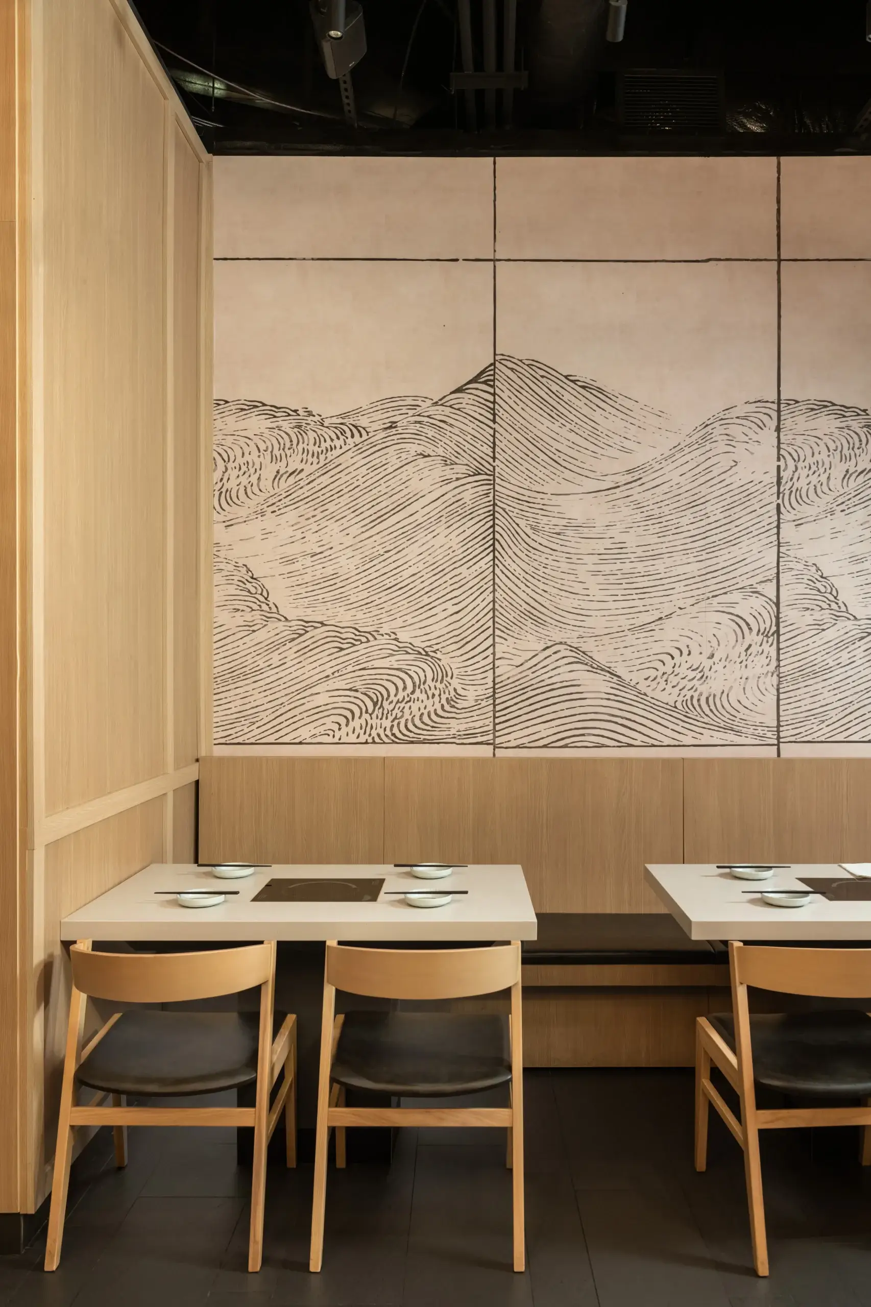 A cozy restaurant corner with light wood paneling, white tables set with chopsticks and bowls, and a large wall mural of flowing, wave-like black lines on beige tiles.