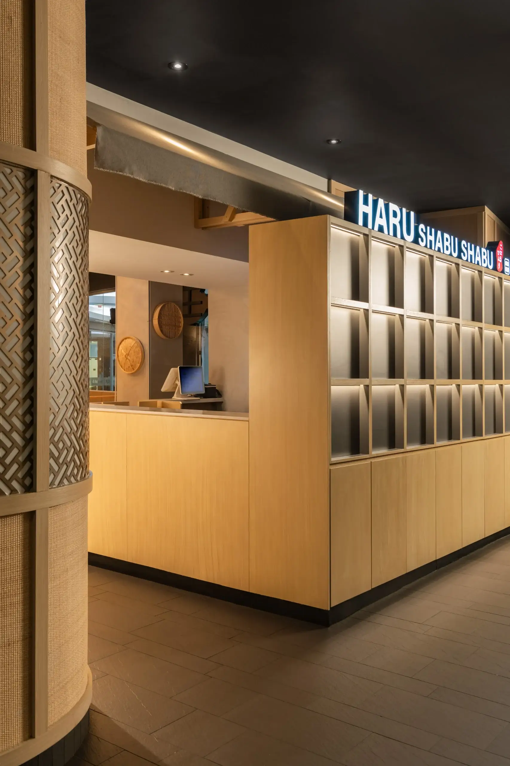 Interior view of a modern dining area with a wooden front desk and a wall of open cubbies behind, illuminated sign reading 'HARU SHABU SHABU' above. Sculpted column with woven patterns on left and warm ambient lighting.