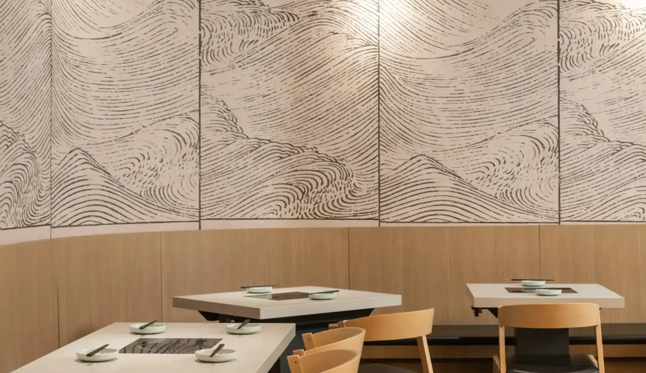 A modern restaurant interior featuring light wood chairs and white tables. The back wall displays a large monochrome wave-pattern mural segmented by panels, with a dark ceiling and track lighting.