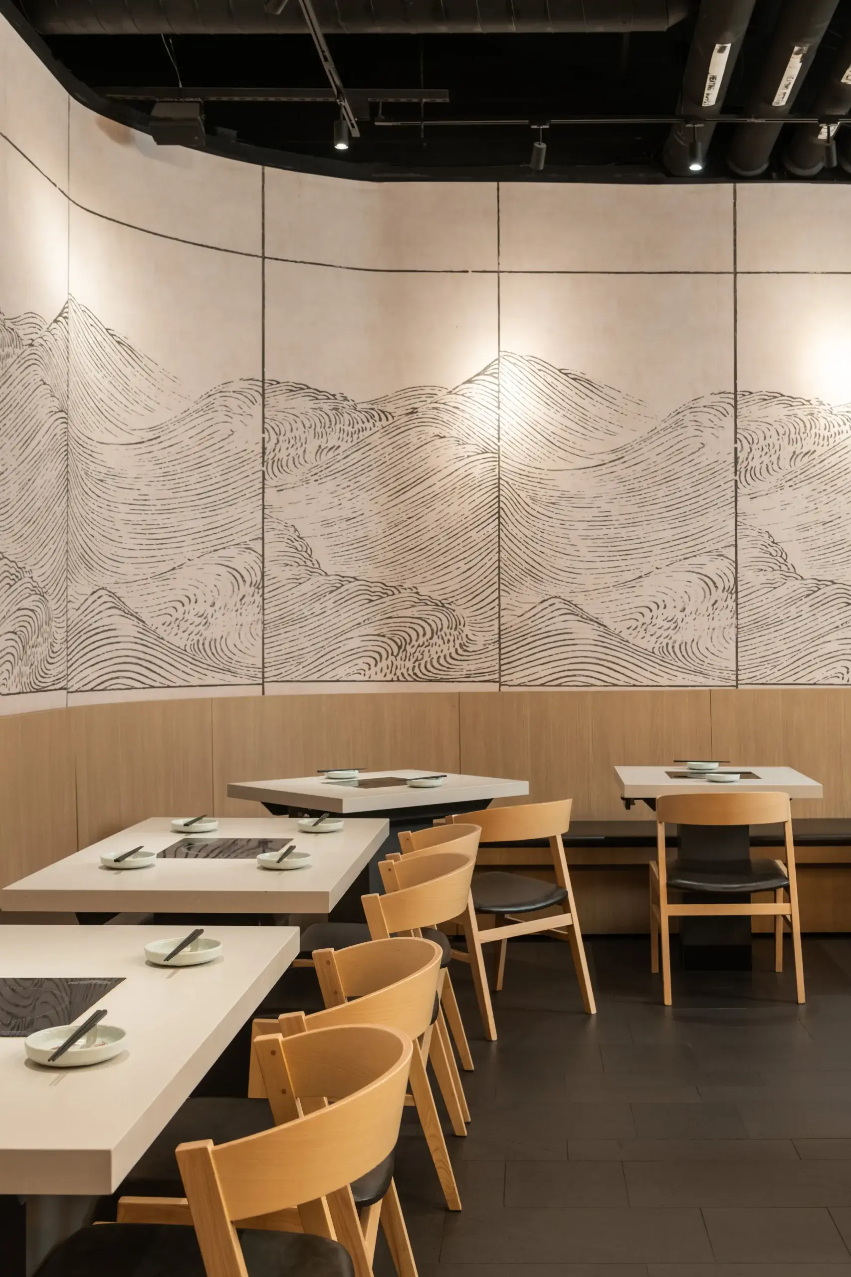 A modern restaurant interior featuring light wood chairs and white tables. The back wall displays a large monochrome wave-pattern mural segmented by panels, with a dark ceiling and track lighting.