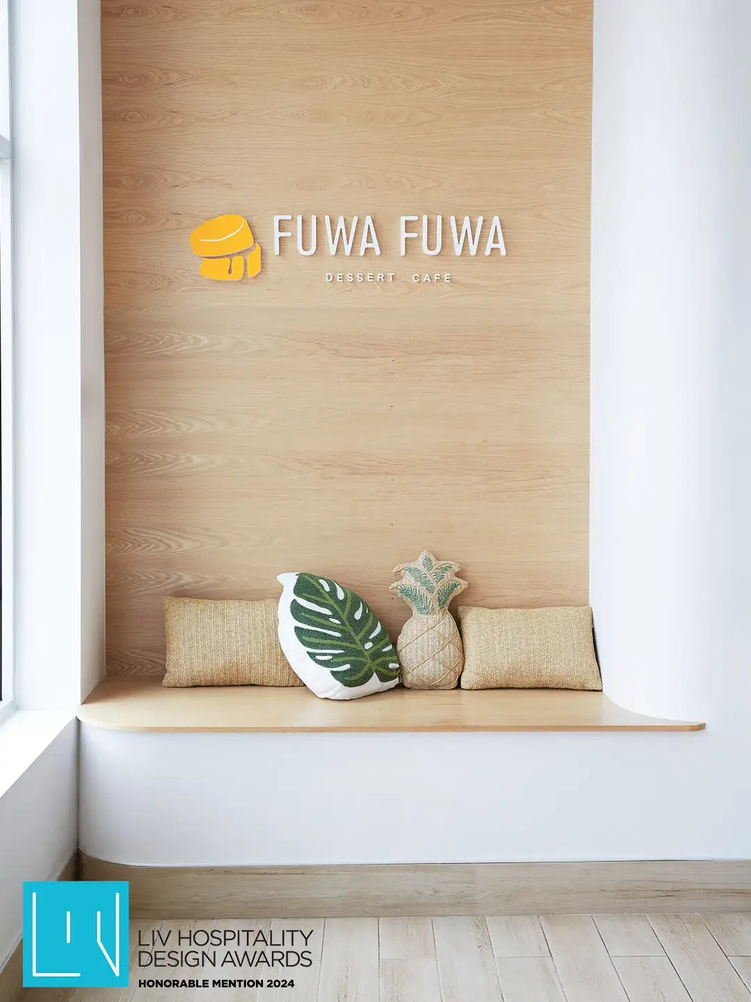 Wood-paneled wall with the sign 'FUWA FUWA DESSERT CAFE' and a small yellow pottery logo. A built-in bench with beige pillows and a decorative leaf-shaped cushion sits beneath, near a window corner.
