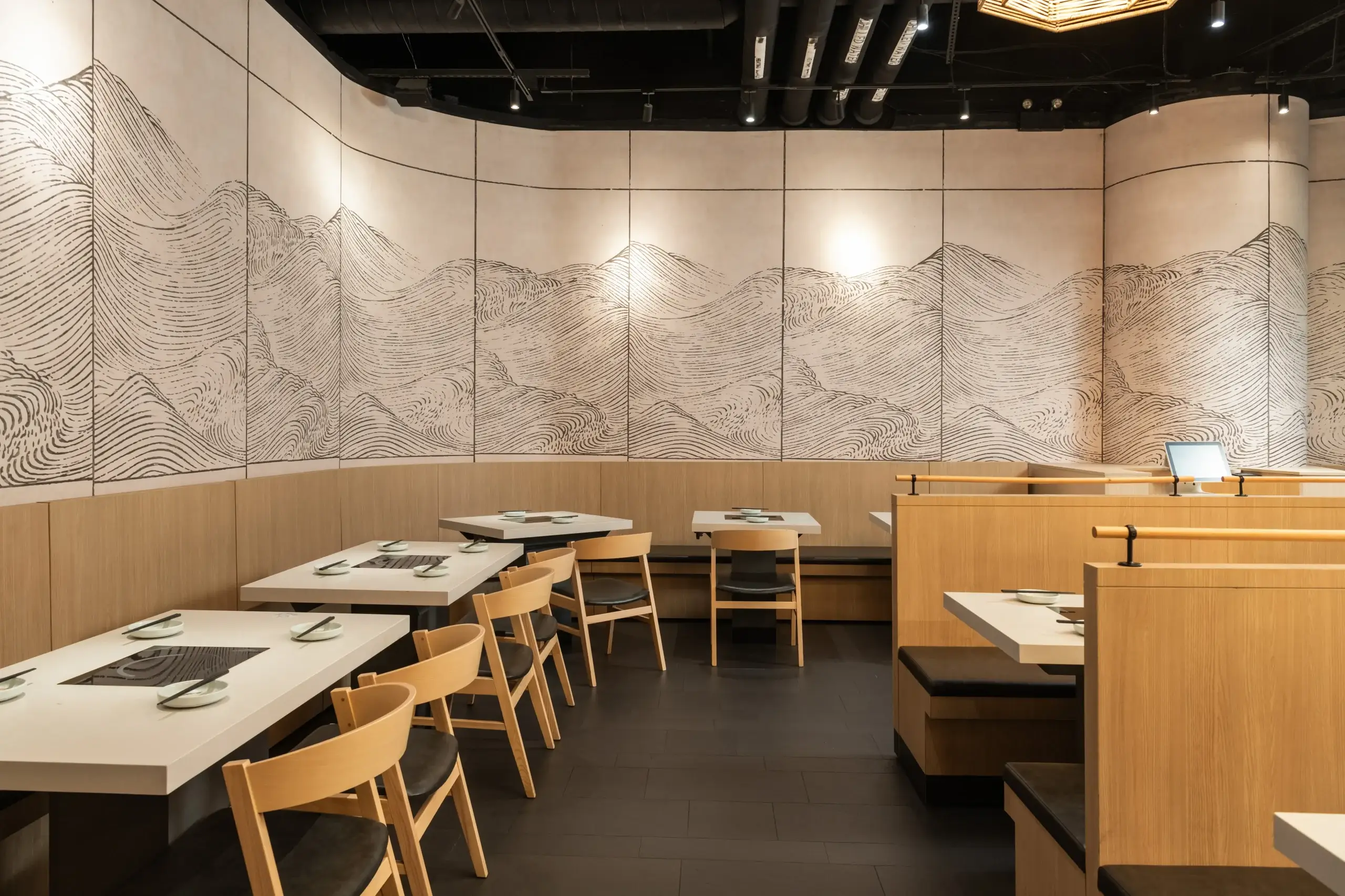 A modern restaurant with light wood seating and white tables arranged along a curved wall. The wall features a large black line art mural resembling stylized mountains, illuminated by ceiling spotlights.