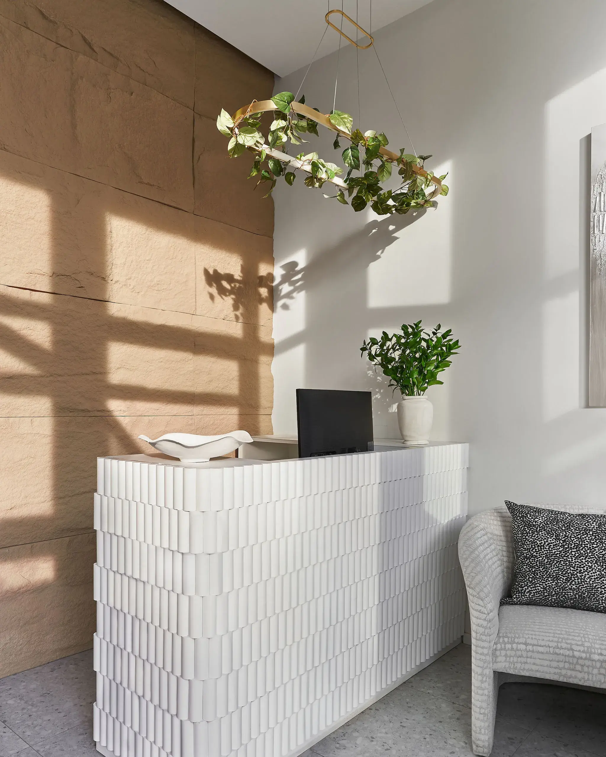 Interior scene featuring a textured white stacked console against a beige stone wall, with a laptop, white bowl, and a potted plant. A hanging circular plant arrangement casts shadows on the wall, and a gray sofa with a patterned cushion sits to the right.