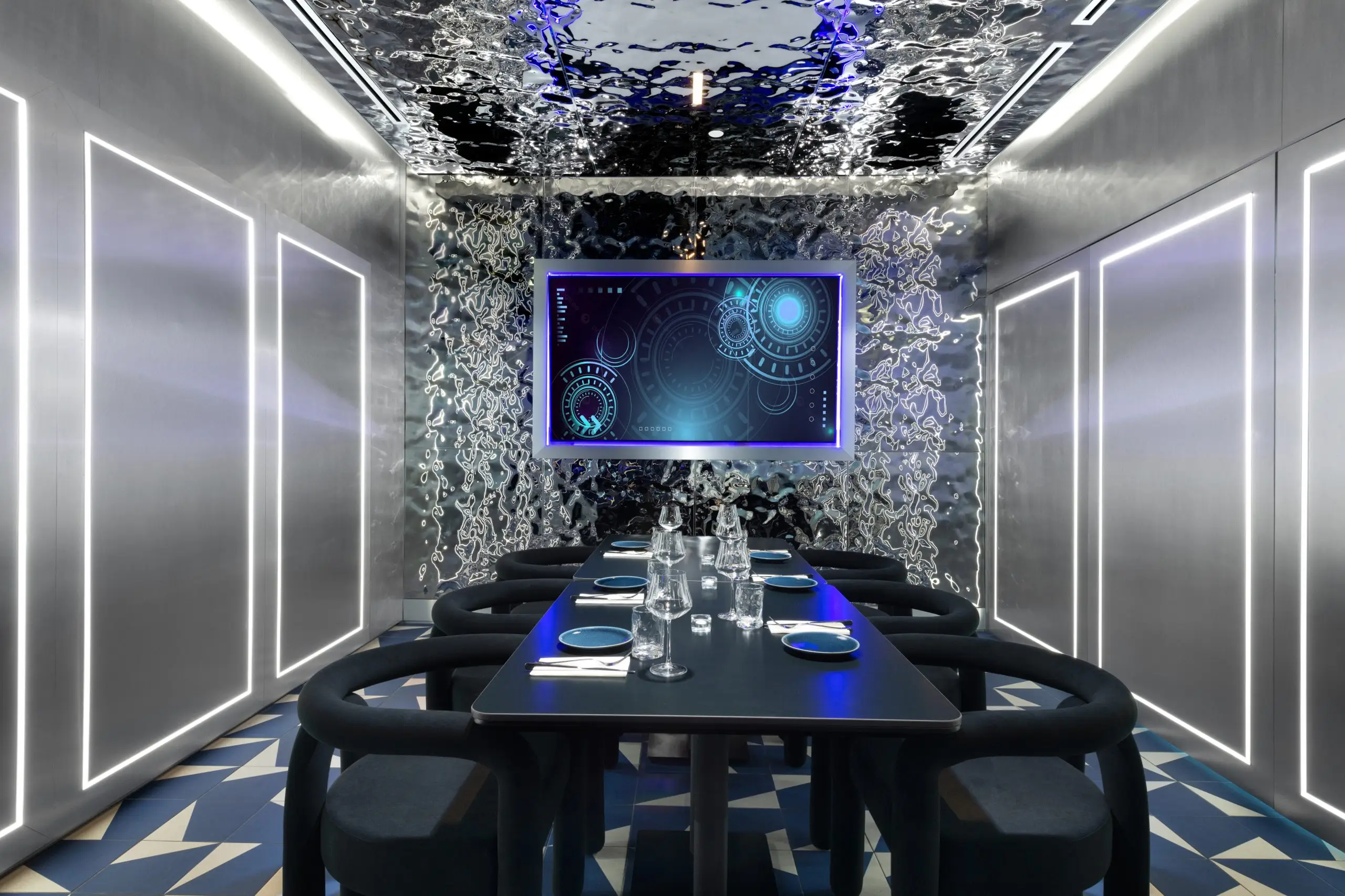 A sleek, high-tech dining room enclosure with metallic walls, illuminated rectangular panels, and a central black table set with plates, glasses, and cutlery. A large screen on the far wall displays blue circular graphics, and the ceiling reflects shimmering patterns.