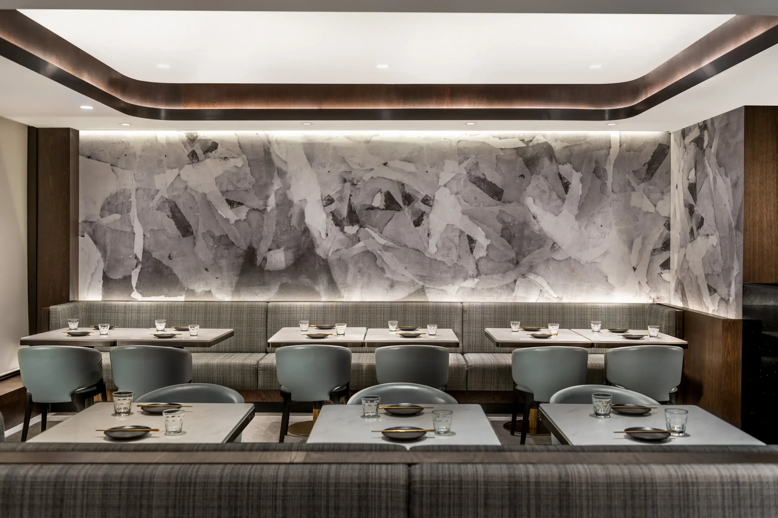 Modern restaurant interior with a long gray marble-patterned wall, curved ceiling lighting, and upholstered banquette seating. Tables are set with plates and glasses, and light gray chairs are arranged around each table.