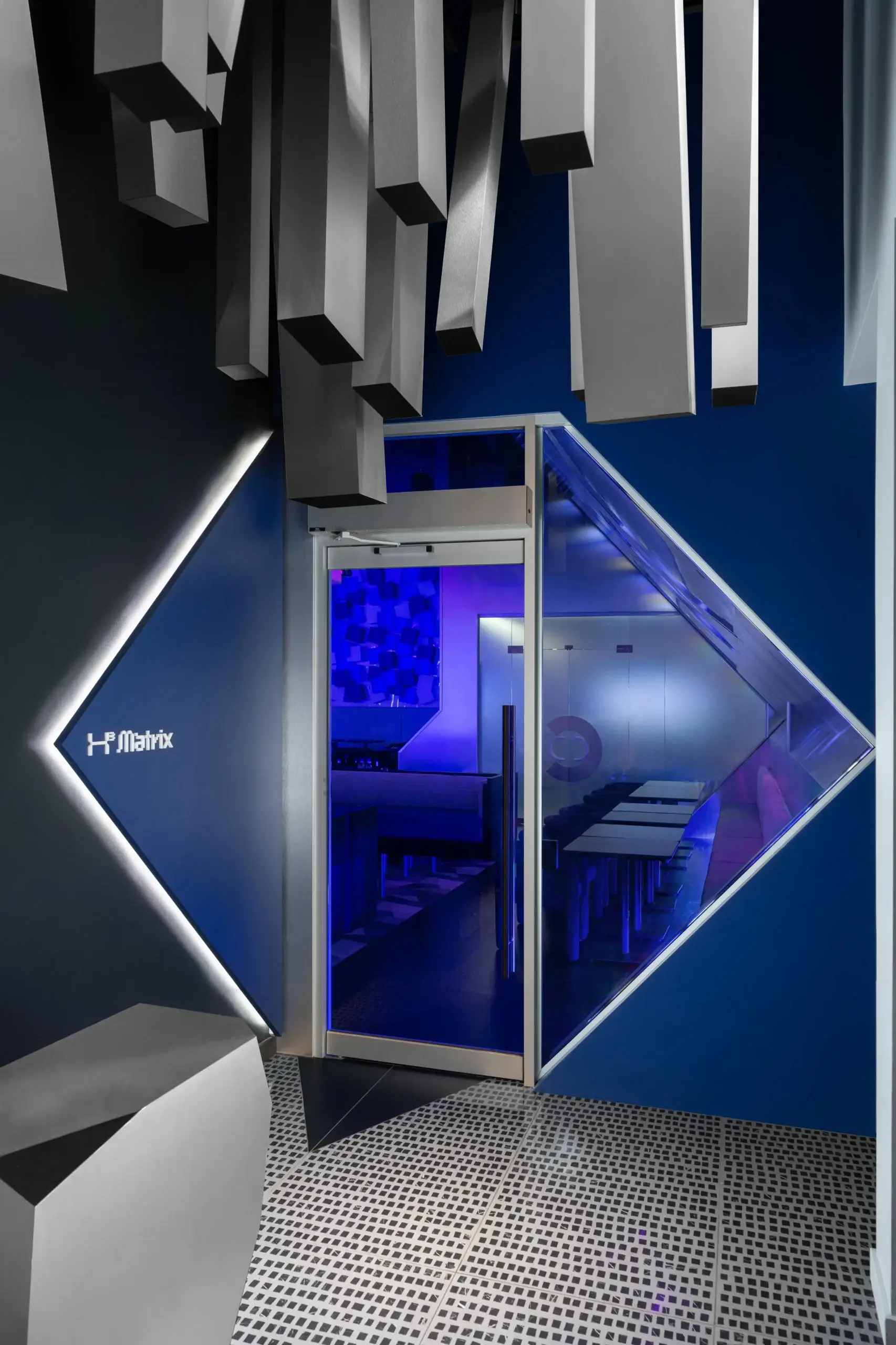 Modern interior with a glass door framed by bright blue walls and angular metallic panels above. A blue LED glow lights a geometric diamond-shaped window or mirror on the wall, and a perforated metal floor leads into a sleek room beyond.