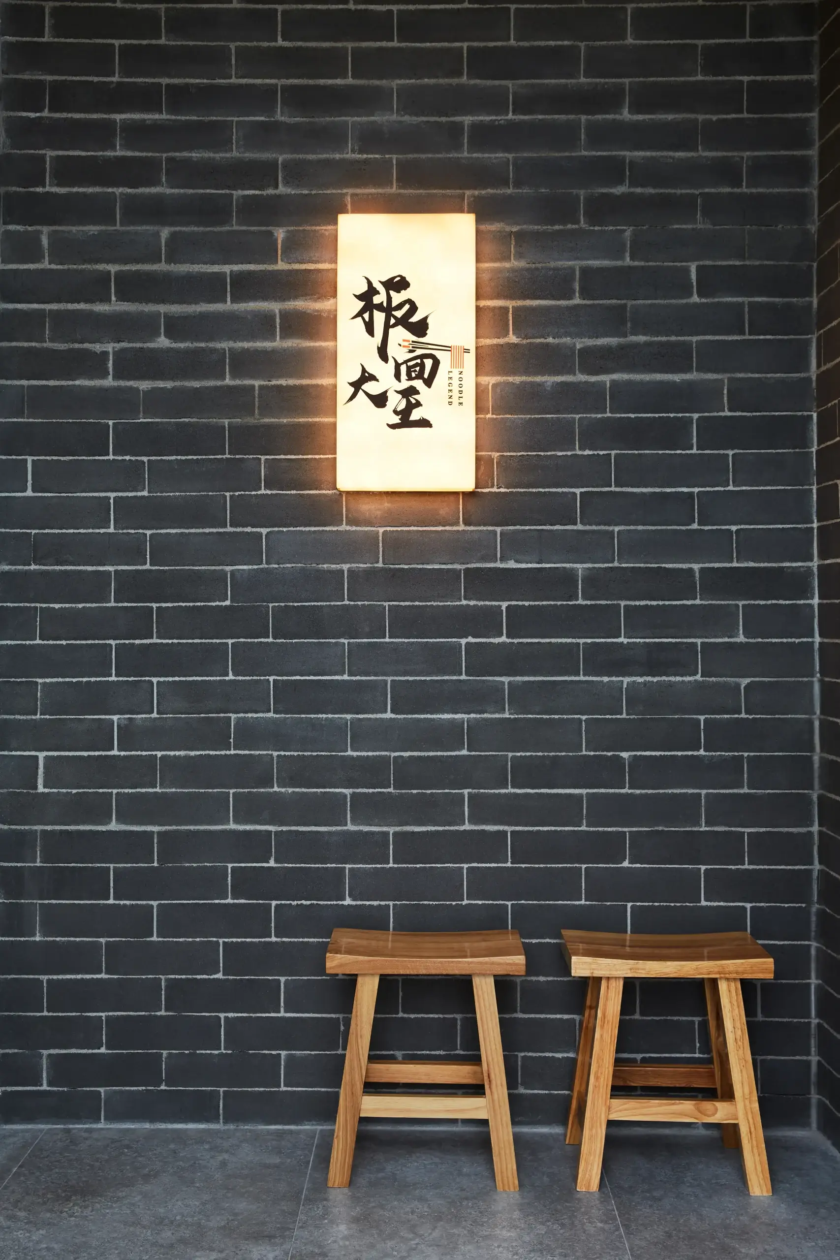 A dark brick wall with a warmly lit rectangular sign featuring Asian characters, centered above two wooden stools with angular legs.