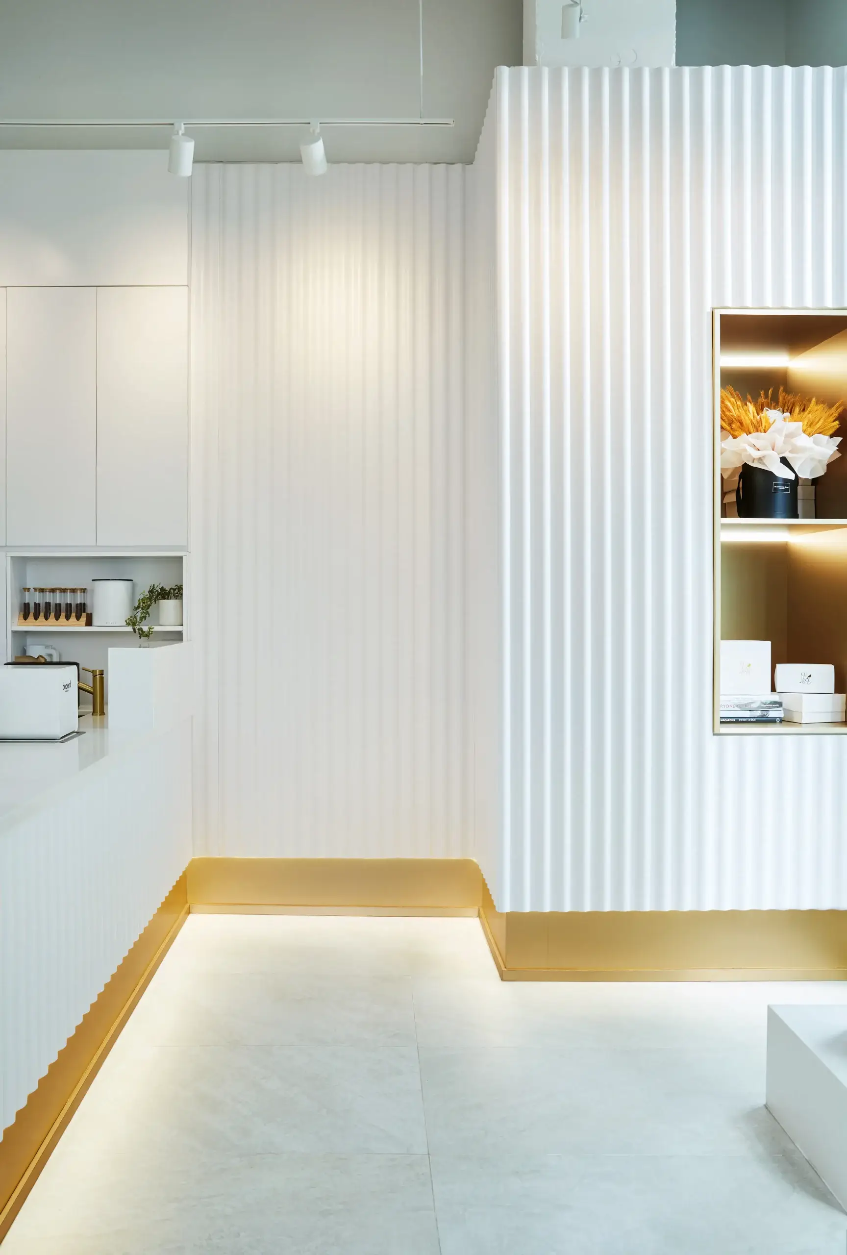 A bright, minimalist interior featuring white corrugated wall panels, a white countertop, and a gold baseboard trim along the floor. Track lighting on the ceiling highlights a clean, contemporary space with a display shelf and subtle decorative items.