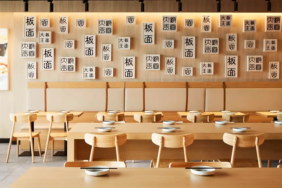 A bright, airy Japanese-style restaurant interior with light wood tables and chairs. The wall is covered with white cards bearing black kanji characters, arranged in a decorative pattern above a long bench seating area. Each table has a small white plate and a pair of chopsticks.