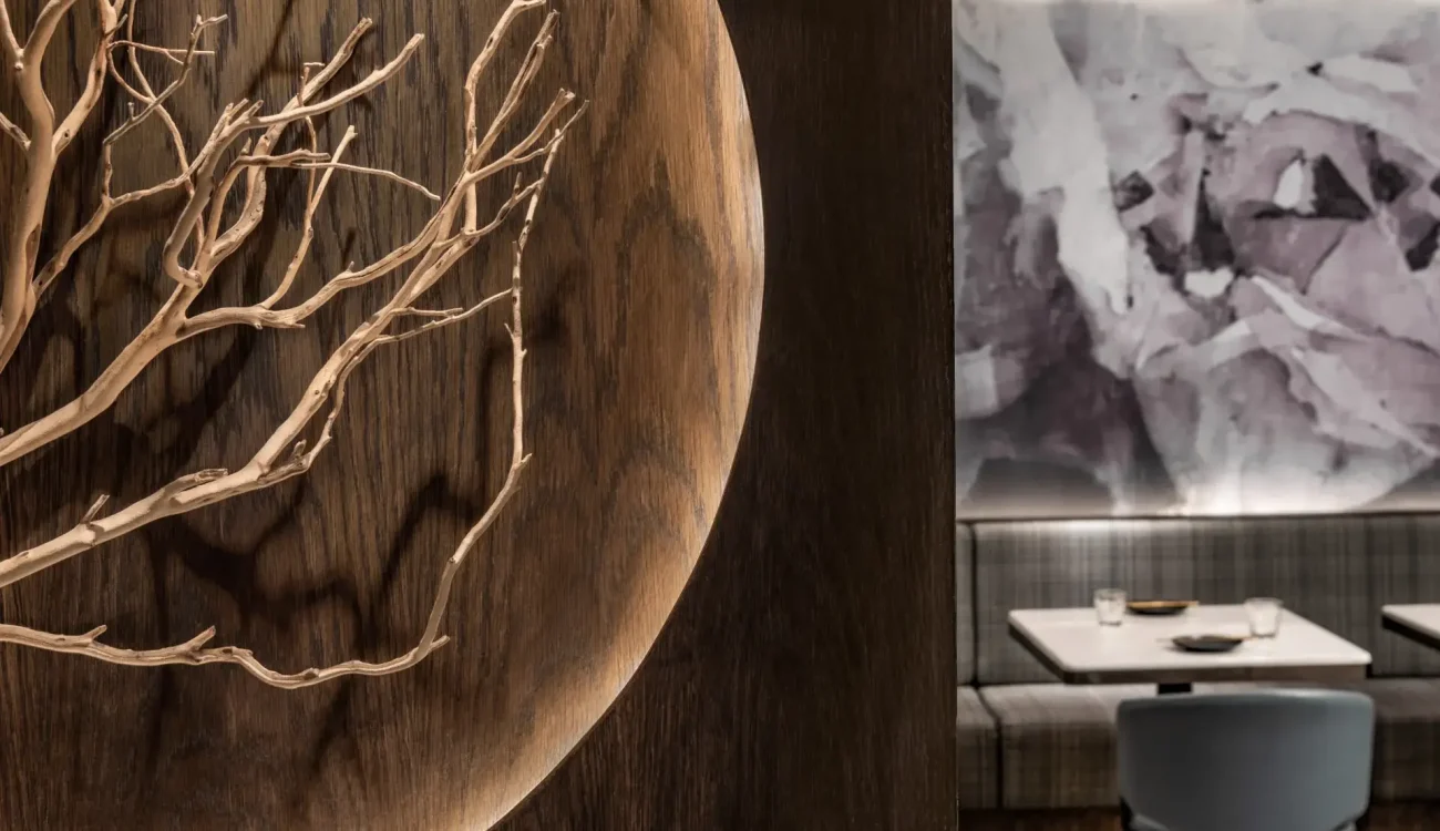 A dark wooden wall features a circular carved inset with light-toned, intricate branch sculpture. In the background, a seating area with gray chairs, white tables, and a marble-patterned wall panel creates a sophisticated dining ambiance.