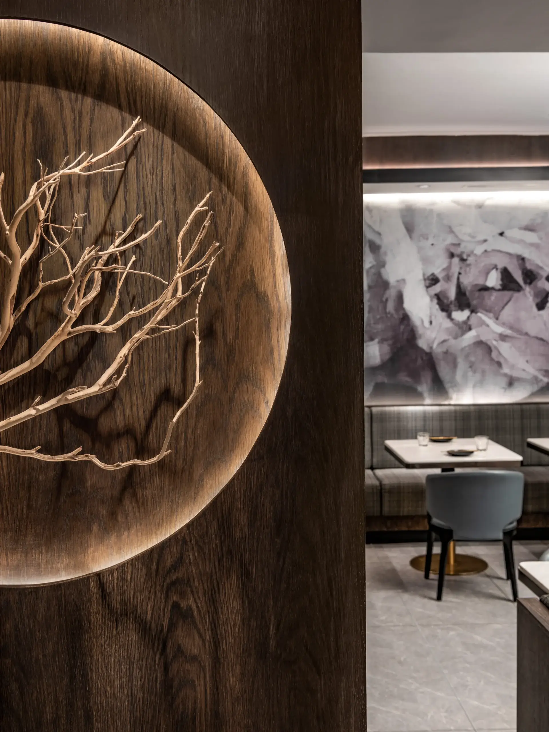 A dark wooden wall features a circular carved inset with light-toned, intricate branch sculpture. In the background, a seating area with gray chairs, white tables, and a marble-patterned wall panel creates a sophisticated dining ambiance.