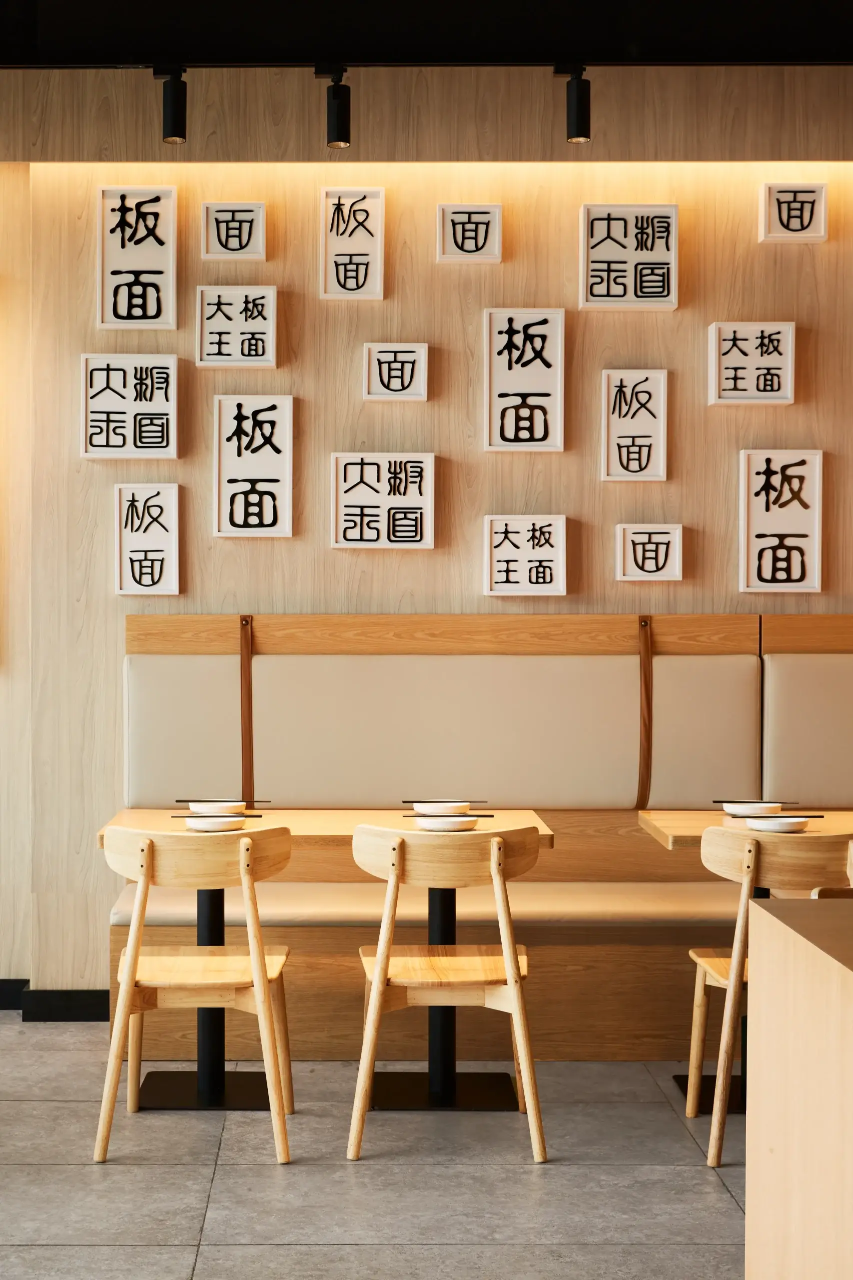 Wooden dining area with light beige booth seating and small wooden chairs. The wall behind features a collage of white framed panels with large black Japanese kanji characters. Warm overhead lighting.